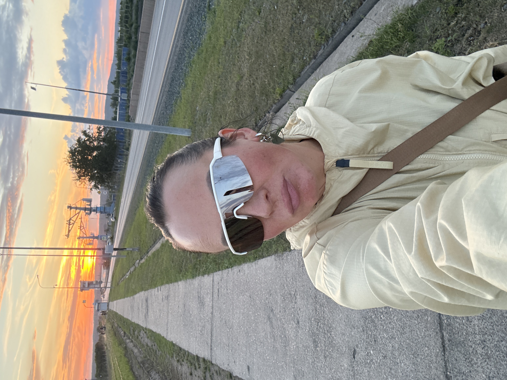 This is a portrait-oriented, slightly tilted, selfie-style photograph taken outdoors during sunset, likely in Chaikovsky, Russia, given the context. The primary subject is a person, viewed from the chest up, wearing a light-colored hooded jacket and large, white, reflective sunglasses. The person has their hair pulled back and is looking towards the camera.
The background shows a paved walkway or path, with grass and gravel on either side. Beyond that, a road or highway is visible, with a median strip. The sky is filled with dramatic sunset colors – oranges, yellows, and pinks – with scattered clouds and the silhouette of distant trees and possibly industrial structures or cranes. The setting appears to be along a road, possibly near an industrial area or transportation hub, given the presence of tall poles and what look like overhead gantries in the distance.
The time of day is clearly sunset, indicated by the vibrant colors in the sky and the low angle of the sun, which is visible in the distance. The weather appears clear and calm, suitable for an outdoor activity. The overall mood conveyed is relaxed and perhaps reflective, given the scenic backdrop. There is no discernible text visible in the image.