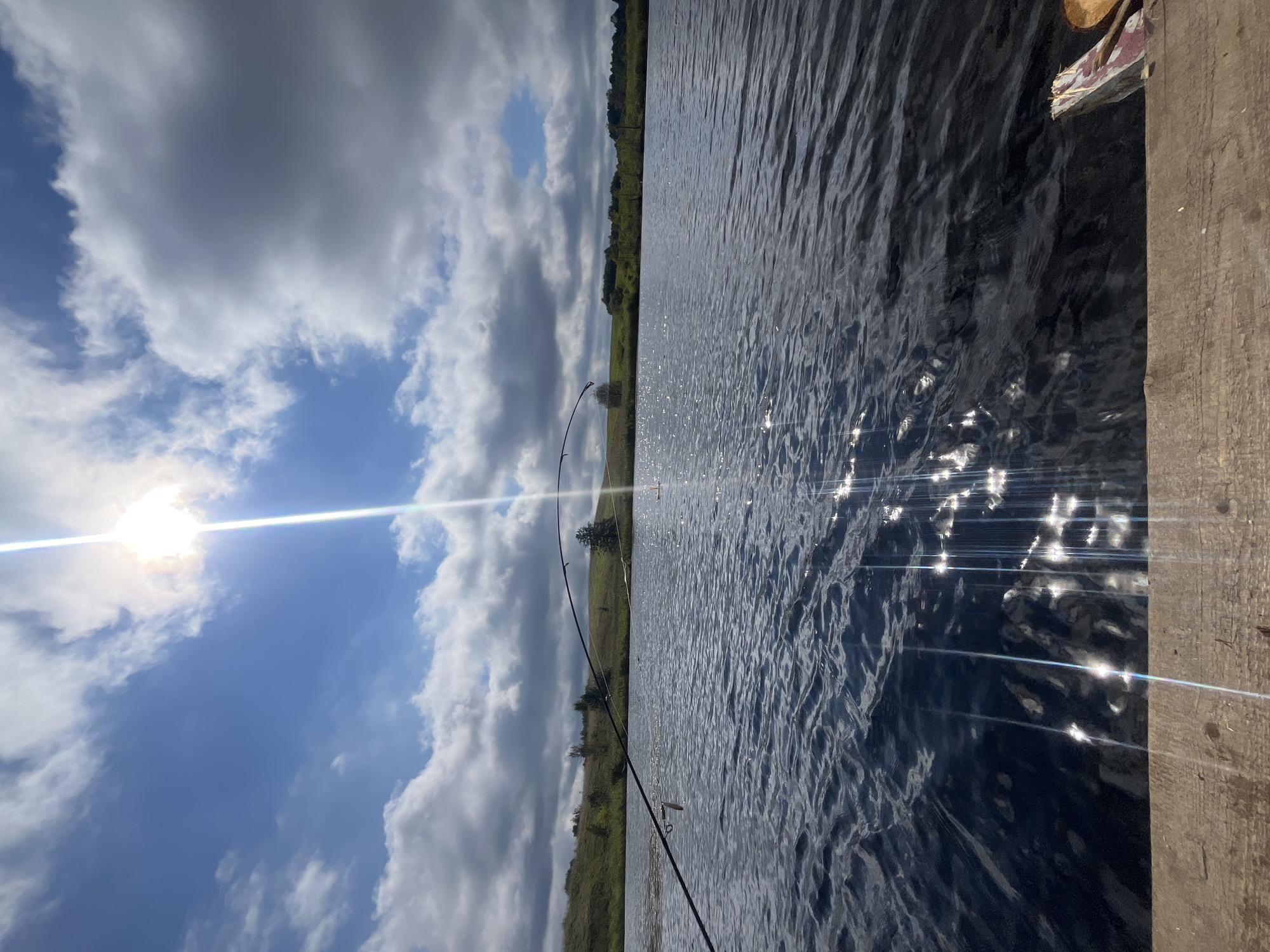 The image shows a lake in the vicinity of Dunilavichy rural council, Belarus, on a sunny day with scattered clouds. The viewpoint is from a wooden surface at the edge of the water. Two fishing rods are visible, one in the foreground arching over the water and another further back. The lake's surface is rippled and reflects the bright sunlight, creating streaks of light. The background features a gently rolling, green hillside dotted with trees. The sky is blue with dramatic white and grey clouds, and the sun is high and bright, emitting a strong glare.
