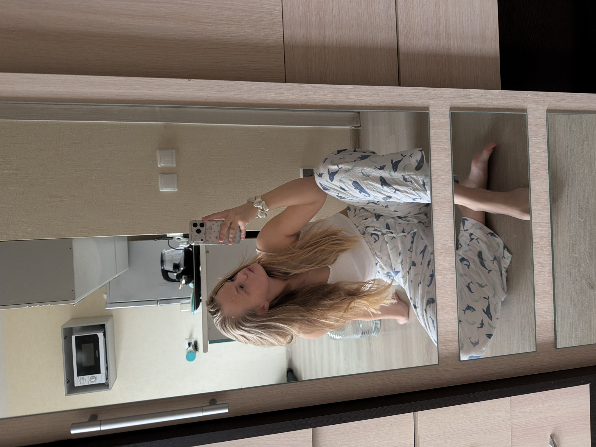A young woman is taking a selfie in a mirror. She is wearing a white top and pajamas with a whale pattern. The mirror reflects a kitchen area with a microwave, cabinets, and light switches. The mirror has a multi-paneled design, with individual mirrored squares creating a frame for the overall reflection. The scene appears to be inside an apartment or home, potentially located in Izhevsk, Russia, based on details such as the interior design.