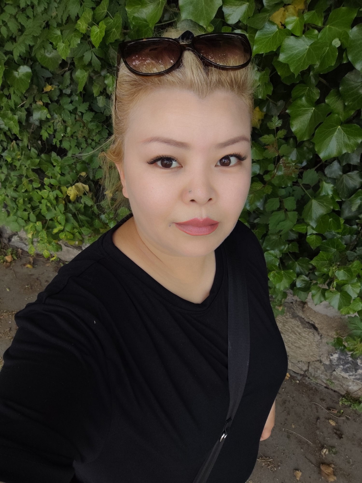A person is standing outside in Sarıyer, Turkey, posing for a selfie-style photograph. The person has light blond hair, brown eyes, and fair skin. They are wearing a black t-shirt and have sunglasses perched on their head. A thin strap, likely from a bag, crosses their body. The background consists of dense green foliage, possibly ivy, growing on a wall or fence. The ground is visible in the lower portion of the frame, showing a paved or concrete surface.