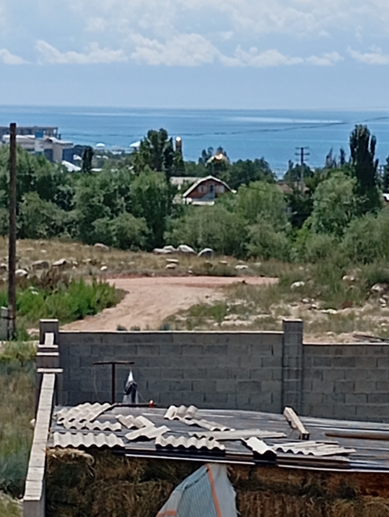 The image depicts a partially constructed or dilapidated rural scene overlooking a body of water, likely Lake Issyk-Kul, given the context of Cholpon-Ata, Kyrgyzstan. In the foreground, a low brick wall runs horizontally across the frame, partially obscuring a structure covered with corrugated metal roofing. This structure appears to be built on top of what looks like bales of hay or straw, with some exposed construction materials on the roof, including wooden planks and more corrugated metal sheets. A thin, weathered wooden post stands to the left, a utility pole with wires is visible in the mid-ground to the right, and a cluster of trees and vegetation dominates the mid-ground. Beyond the vegetation, a small building with a red roof is visible, and further in the distance, a larger body of water stretches to the horizon under a cloudy sky. The water's edge shows some distant buildings and industrial structures. The overall impression is one of a developing or unfinished area, common in the outskirts of towns like Cholpon-Ata.