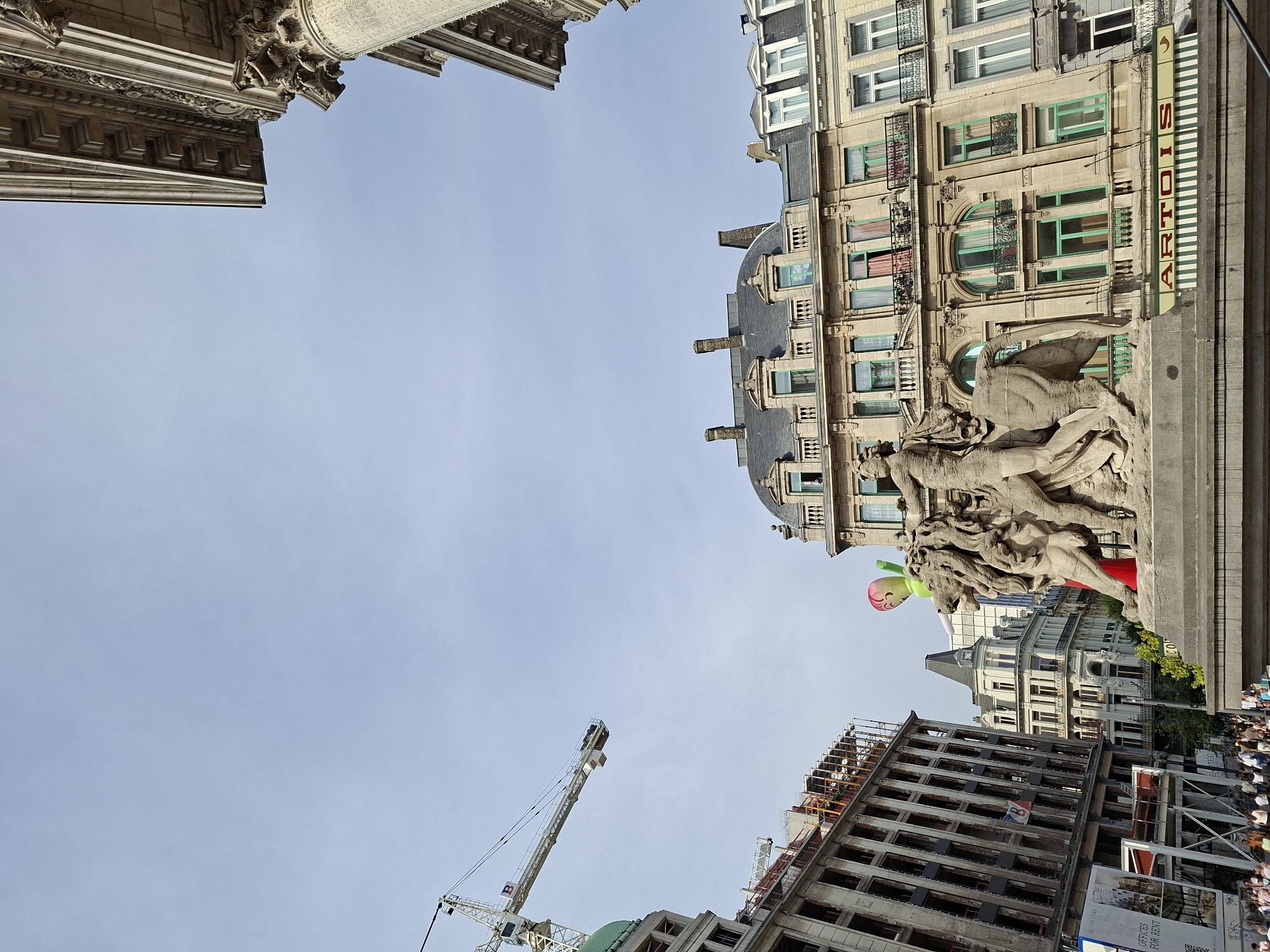 A street-level, eye-level, medium shot captures a busy urban scene in Brussels, Belgium. In the foreground, a large stone sculpture depicts multiple figures, including a man and what appears to be a lion, in a dynamic pose. Behind the sculpture, a multi-story building with ornate architecture and green-framed windows dominates the mid-ground. Several other buildings of varying architectural styles, including one under construction with scaffolding, line the street. A construction crane is visible in the sky to the left. A crowd of pedestrians walks along the sidewalk in the lower portion of the frame, suggesting a public space. The sky is clear and pale blue. A sign with the name "ARTOIS" is visible on the building behind the sculpture.