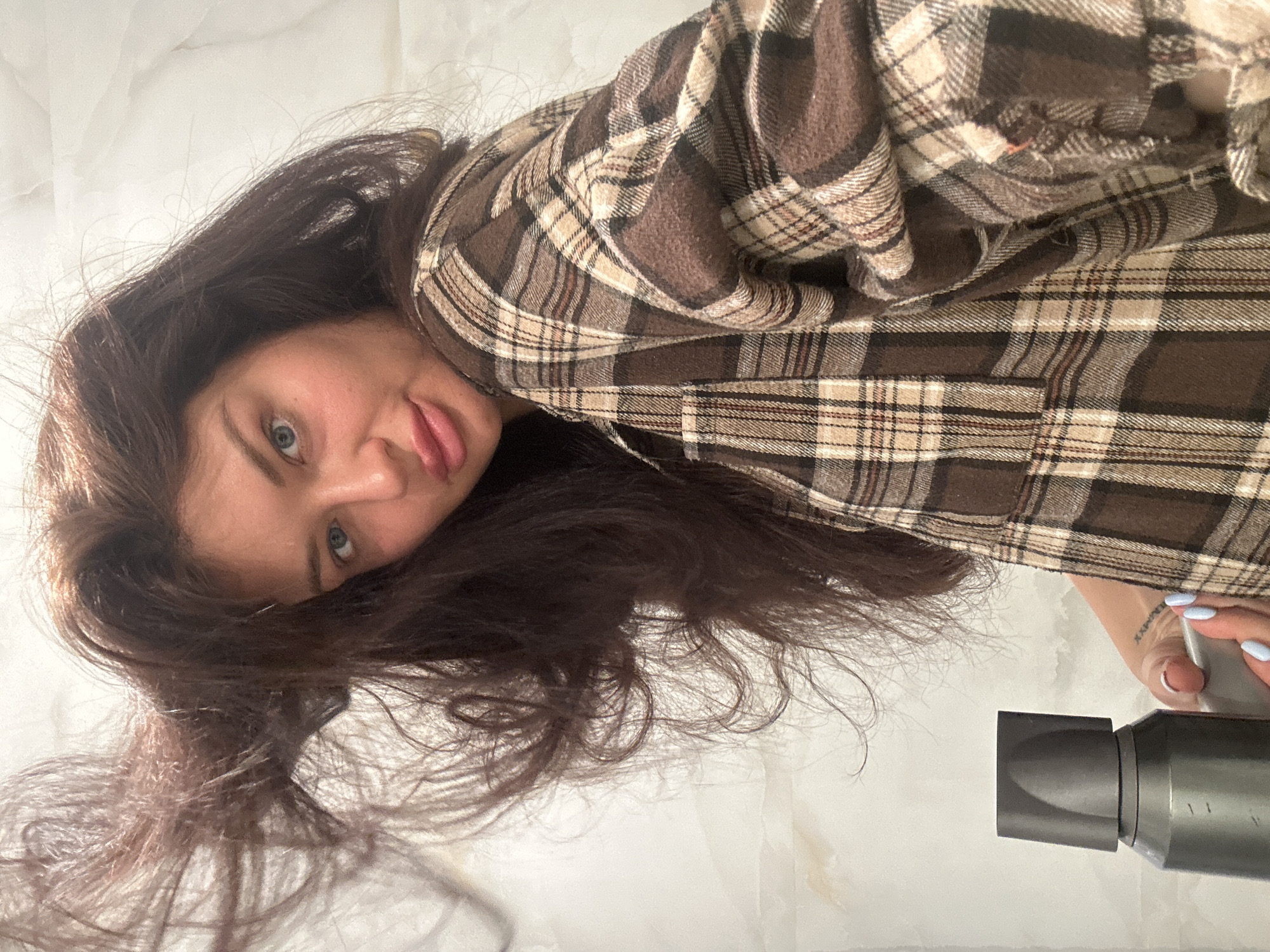 The image is an upward-angled selfie of a woman with dark brown hair, light blue eyes, and fair skin. She is wearing a brown and tan plaid flannel shirt. Her hair is loose and somewhat messy. She is sticking out her tongue slightly. In the lower right corner, part of a gray hairdryer is visible. A portion of her hand, with pale blue nail polish, is holding the hairdryer. The background appears to be a white or light-colored marble or tiled surface. There is no indication of the city of Khimki, Russia, or any specific geographic location within the image.