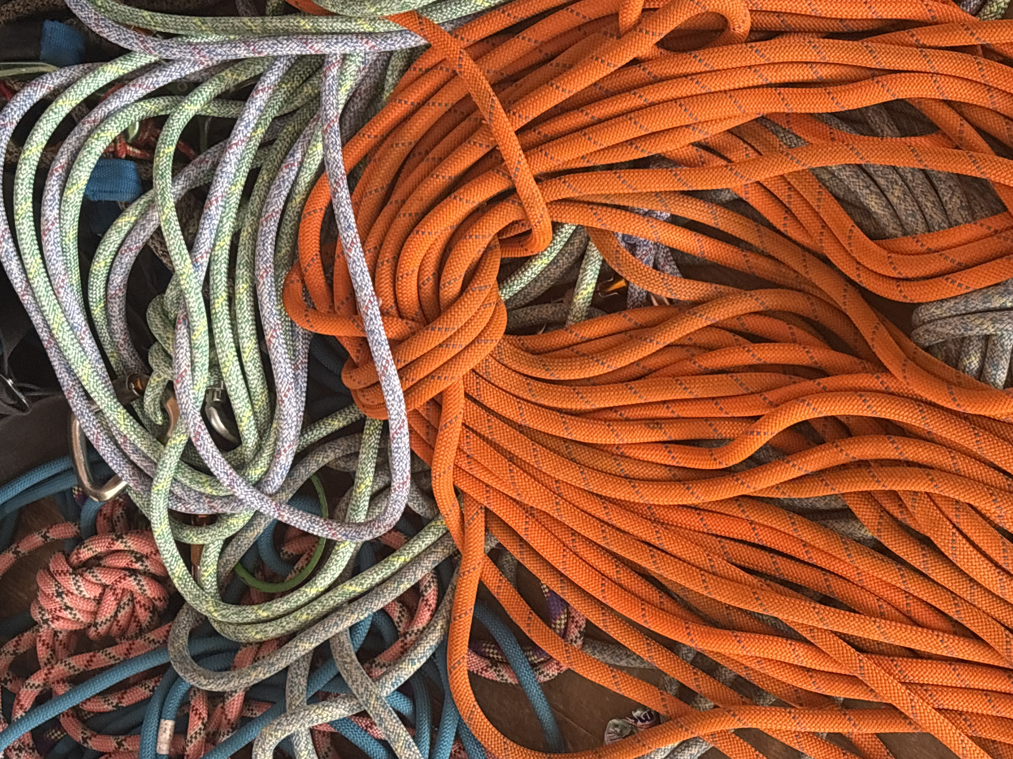 The scene, located in the city of Tayshet, Russia, presents a dense, overhead view of numerous tangled climbing ropes. A prominent, bright orange rope with a black speckled pattern dominates the foreground and midground. Intertwined within this mass are several other ropes of varying colors, including light green, light blue, pink, gray, and purple, all featuring a braided or woven sheath texture with distinct flecking patterns. A silver-colored metallic carabiner is partially visible amongst the lighter colored ropes in the upper left quadrant. The ropes are irregularly coiled and intermingled, resting on an indistinct dark surface. No individuals or specific environmental markers beyond the ropes are discernible.