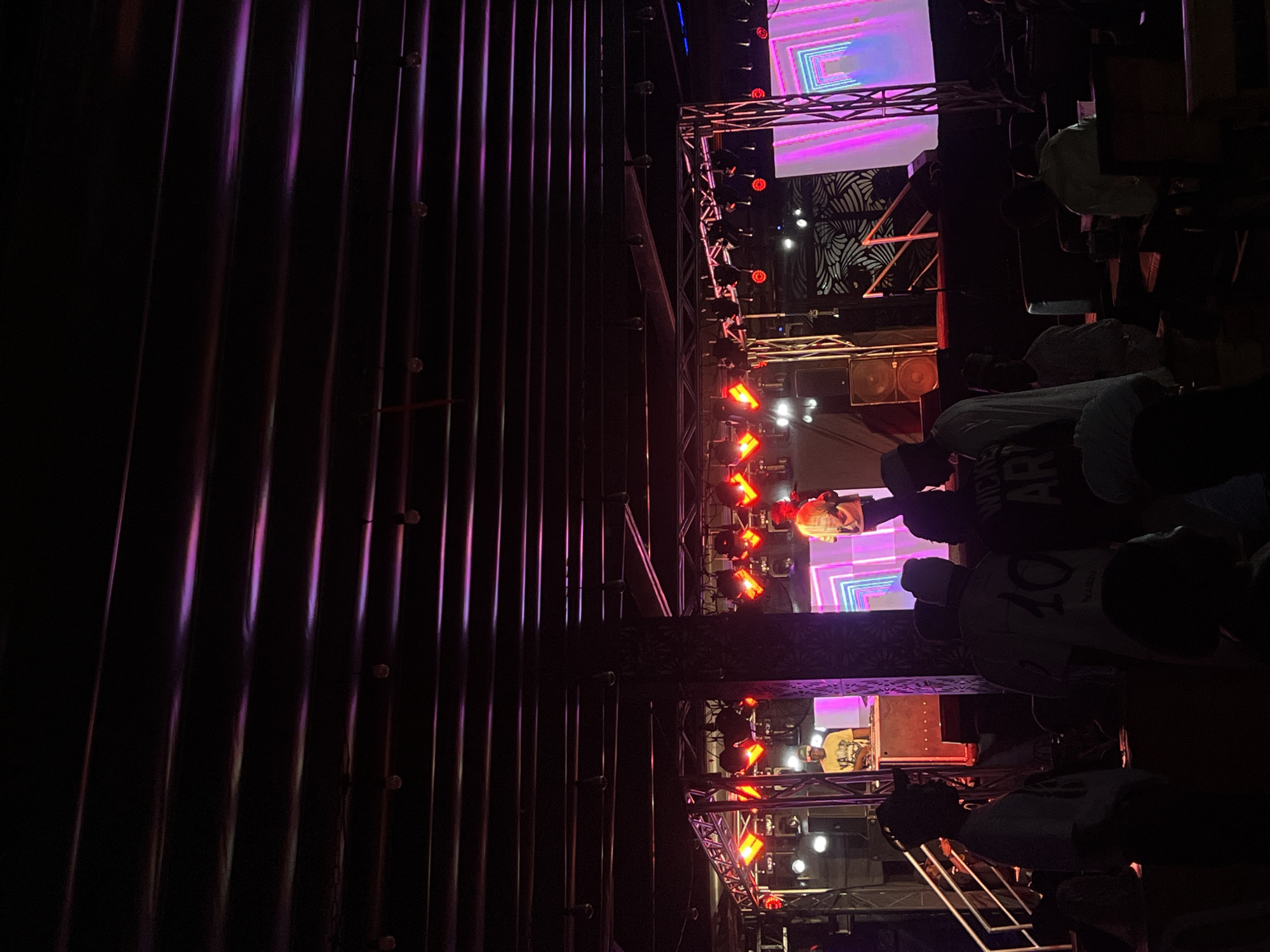 The image depicts a music performance in Howayi, Nigeria, at night. A stage is set up with lighting rigs and a video screen displaying abstract patterns. Several people are visible in the foreground, appearing to be audience members watching the performance. The environment seems to be a venue or open-air space, with metal paneling visible on one side.