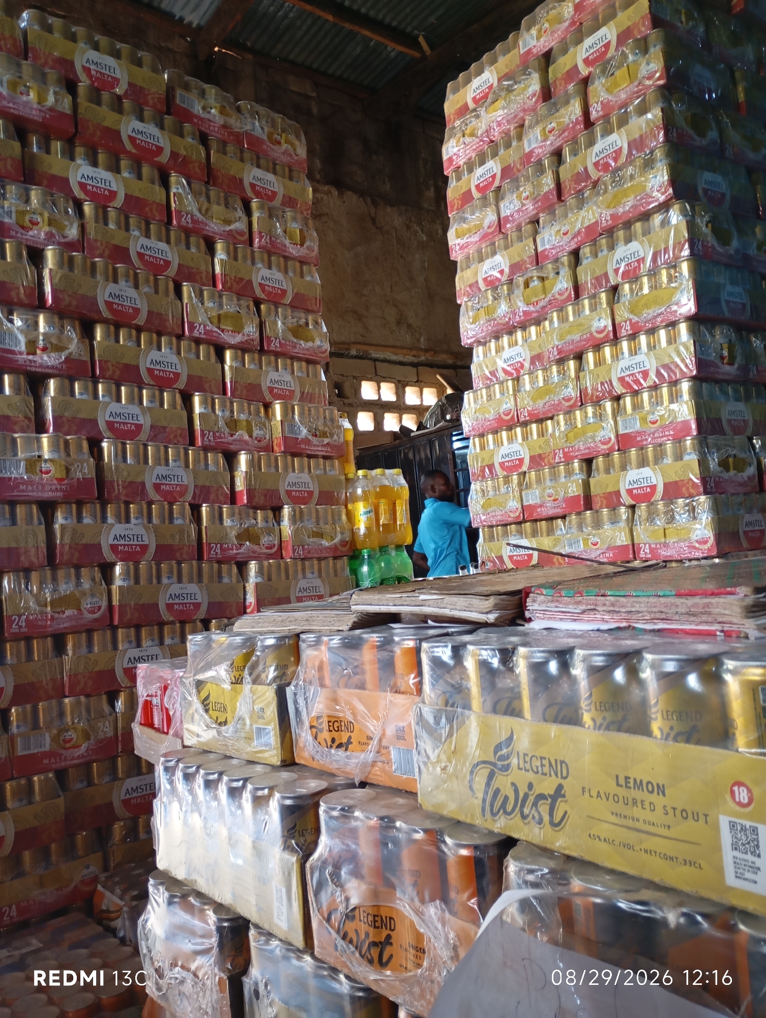 An interior shot shows a warehouse packed with cases of Amstel Malta, stacked high. The cases are wrapped in plastic, and each contains 24 cans. The lighting is dim, with sunlight filtering through gaps in the corrugated metal roof. A man in a blue shirt stands near the center of the image, facing away from the camera, looking towards the stacks. In the foreground, several cases of "Legend Twist Lemon Flavoured Stout" are visible. The date "08/29/2026 12:16" and "REDMI 13C" are watermarked on the image.