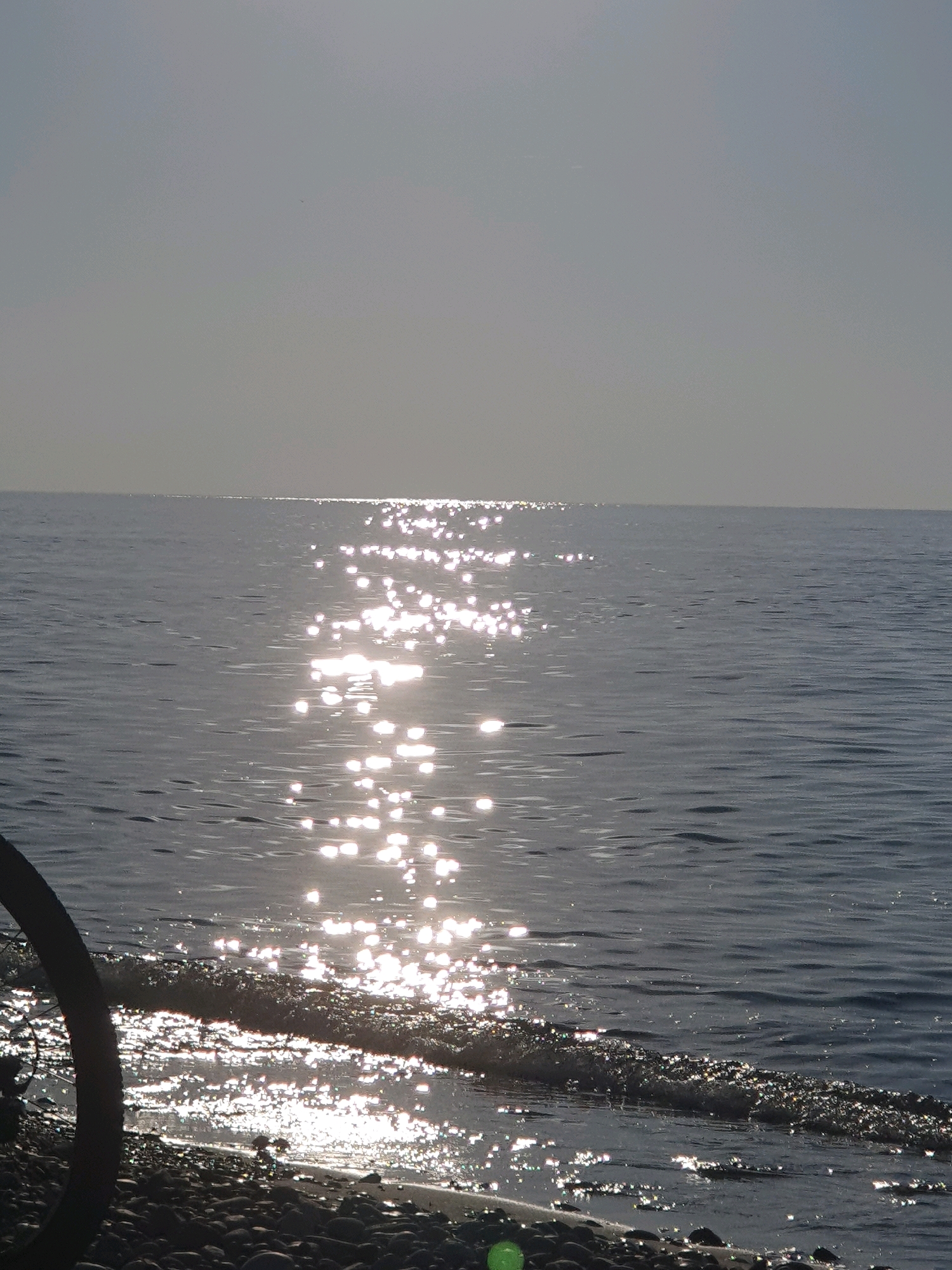The image depicts a serene view of the Black Sea at Batumi, Georgia. The sun's reflection shimmers across the water, creating a dazzling path of light that stretches towards the horizon. The sky is a soft, hazy gray, blending seamlessly with the distant sea. In the foreground, a pebbly shore is visible, with gentle waves lapping at the edge of the water. A dark, curved object, possibly part of a railing or structure, is partially visible on the left side of the frame, grounding the scene. The overall impression is one of peaceful solitude and natural beauty, characteristic of the coastal landscapes found in Batumi.