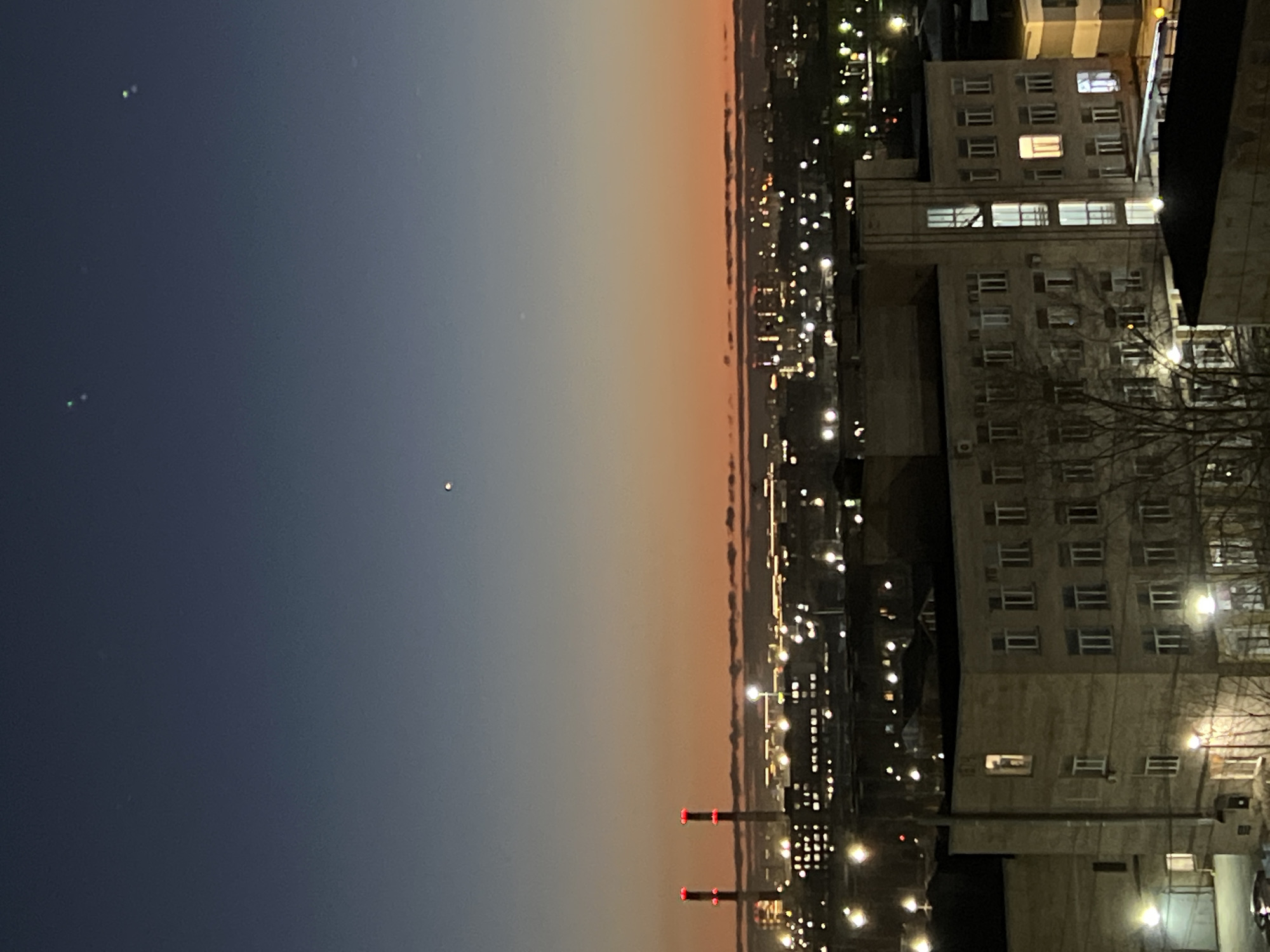 The image depicts a nighttime cityscape, likely taken from an elevated vantage point in Saint Petersburg, Russia. The sky transitions from a dark blue on the left to a warmer orange hue near the horizon line, indicative of either sunset or sunrise. Several points of light are visible in the sky, which could be stars or distant celestial bodies.
To the right of the image is a dense urban area characterized by numerous buildings and streetlights. The buildings appear to be older, multi-story structures, with lit windows suggesting residential or commercial occupancy. A power plant is visible with its chimneys.
The environment is dark and urban, typical of a city at night. There are no people, direct actions or interactions depicted in the image.