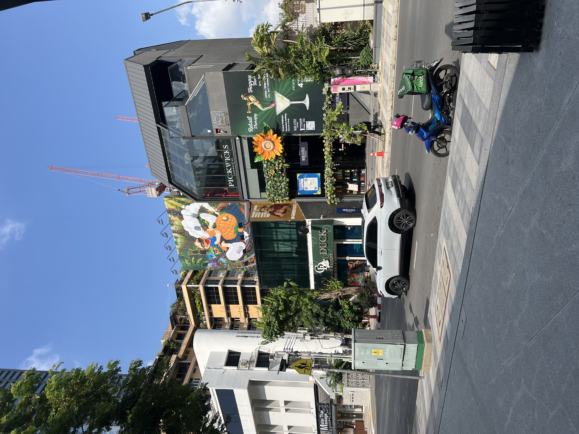 The image depicts a street scene in Bangkok, Thailand, showing a mix of businesses and urban elements. A white car is parked on the street next to a crosswalk, with a blue motorcycle carrying a "Grab" delivery bag parked behind it. The motorcycle driver wears a pink helmet. The buildings include "Pickwicks," identifiable by its prominent signage, "In Green Duck," and "Malai Thai Massage." The Pickwicks' building features a striking sunflower decoration and advertisements for live jazz. There's also a sign for "The Green Duck Cannabis Boutique." Electrical boxes and a pole with street signs are visible on the sidewalk. A crane is seen in the distance, indicating construction activity. The environment suggests a bustling commercial area.