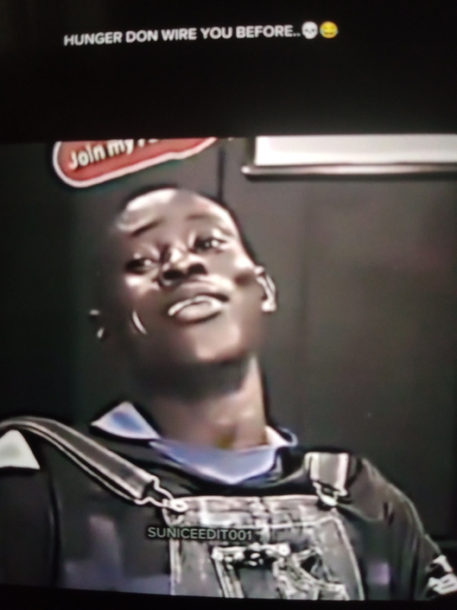 The image shows a close-up shot of a young Black man looking upwards, with a slight smirk. He is wearing dark overalls over a blue collared shirt. Text overlaying the top of the image reads "HUNGER DON WIRE YOU BEFORE..". In the background, a red and white sign with the text "Join my" is partially visible. Another watermark reads "SUNICEEDIT001". The image appears to be a screenshot from a video or social media post, not a photograph taken in Jalingo, Nigeria. The setting is indoors with a dark, indistinct background.