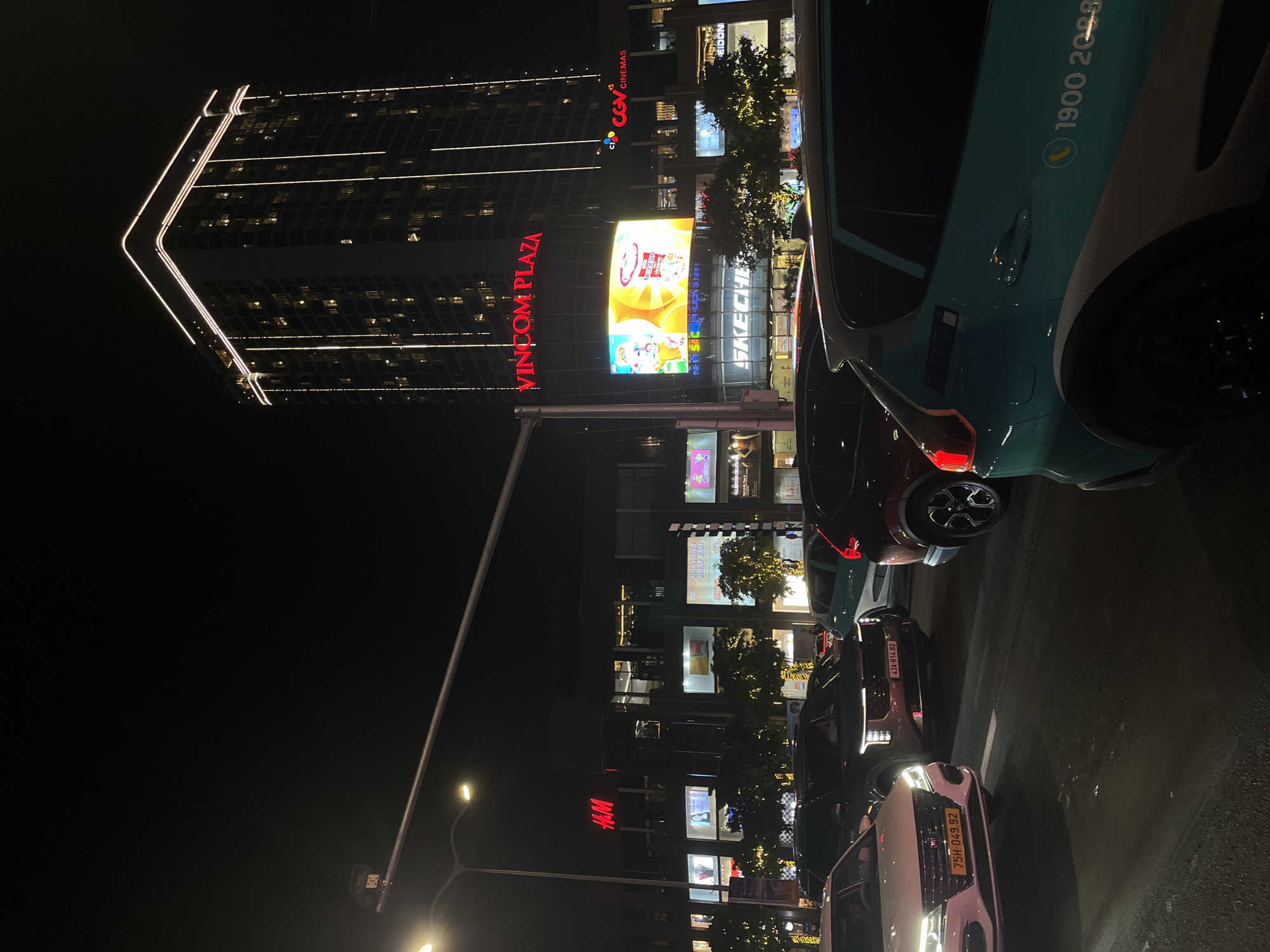 The image captures a nighttime scene in Đà Nẵng, Vietnam, dominated by the illuminated Vincom Plaza, a multi-story shopping center with large storefront signs, including H&M, Skechers, and CGV Cinemas. The building is flanked by a taller, adjacent structure lit with horizontal lines of light. The foreground is filled with parked cars, including a taxi bearing the phone number 1900 2088, partially obscuring the lower portion of the shopping center. Streetlights are visible in the middle ground. License plates on several vehicles are visible.