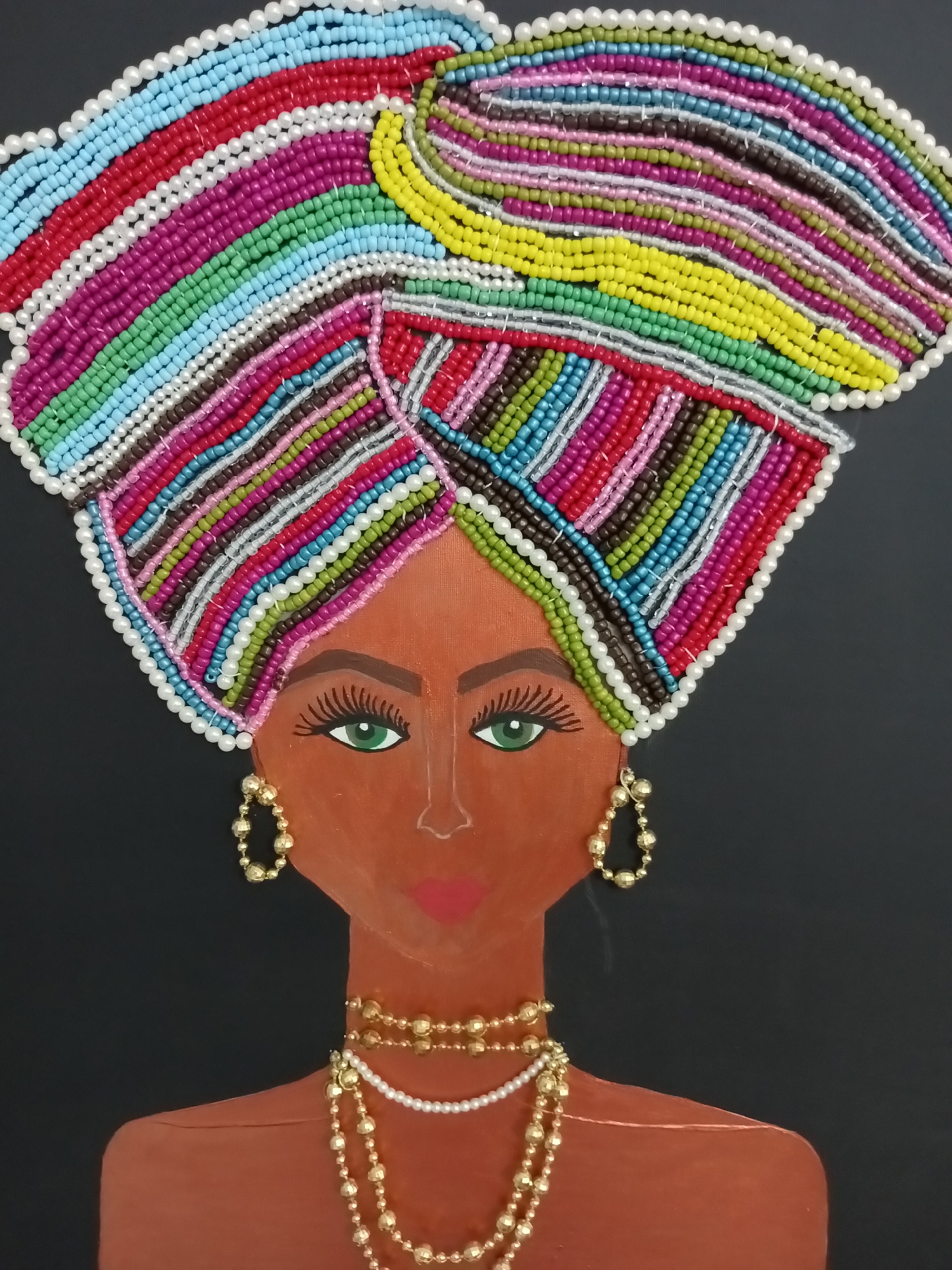 The image depicts a close-up, vertical, eye-level shot of a stylized, handmade portrait of a woman. The background is a solid black color. The woman's face is rendered in a warm, brown tone, with green eyes, dark eyelashes, and pink lips. She is adorned with gold earrings and multiple gold and pearl necklaces. Her head is covered by a large, intricately beaded turban. The turban features a vibrant, multicolored design composed of numerous small beads in shades of red, pink, blue, green, yellow, white, and brown. The beads are arranged in parallel, curved rows, creating a dynamic and eye-catching pattern. The overall presentation is artistic and decorative. There is no indication of the city of Alanya, Turkey, or any specific geographical location within the image.