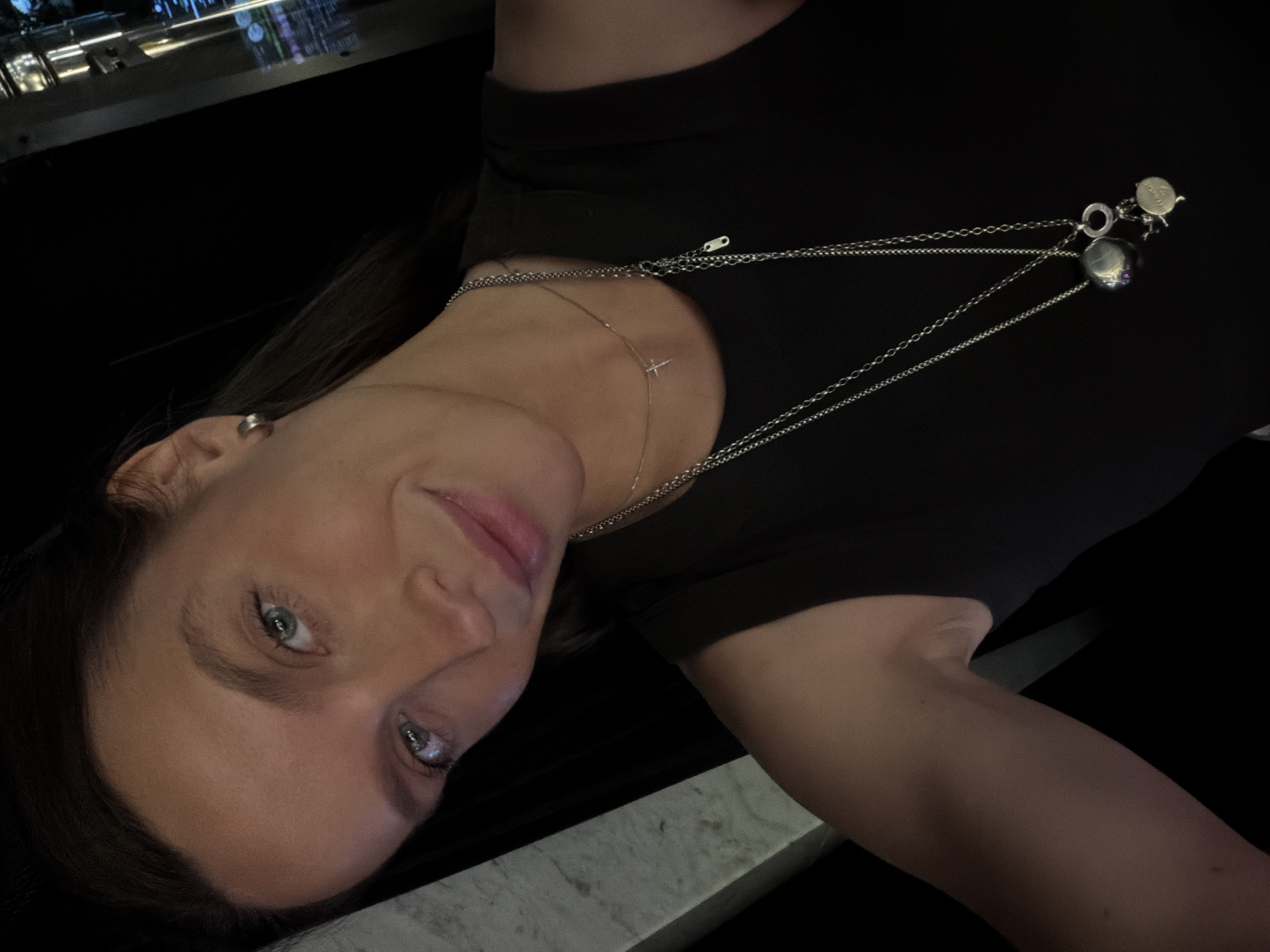 A woman is seen from the chest up, looking at the camera. She has light blue eyes and light brown hair. She is wearing a black sleeveless shirt and two silver necklaces. One necklace has a small cross pendant, and the other has two charms. She has a silver hoop earring in her left ear. The background is dark, with some blurred lights and shapes that suggest an indoor environment, possibly a bar or restaurant in Budva, Montenegro, with a sleek, modern design.