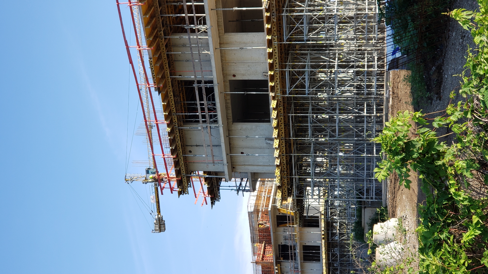 A construction site is depicted in Prešov, Slovakia, under a clear blue sky. The main subjects are unfinished concrete buildings, characterized by visible floors, structural elements, and scaffolding. A tower crane, yellow with a red boom, is positioned in the upper left quadrant, extending over the buildings. Workers are visible on the upper levels of the construction, engaged in work. In the foreground, lush green foliage and some concrete structures are present. The scene captures an active construction project in progress.
