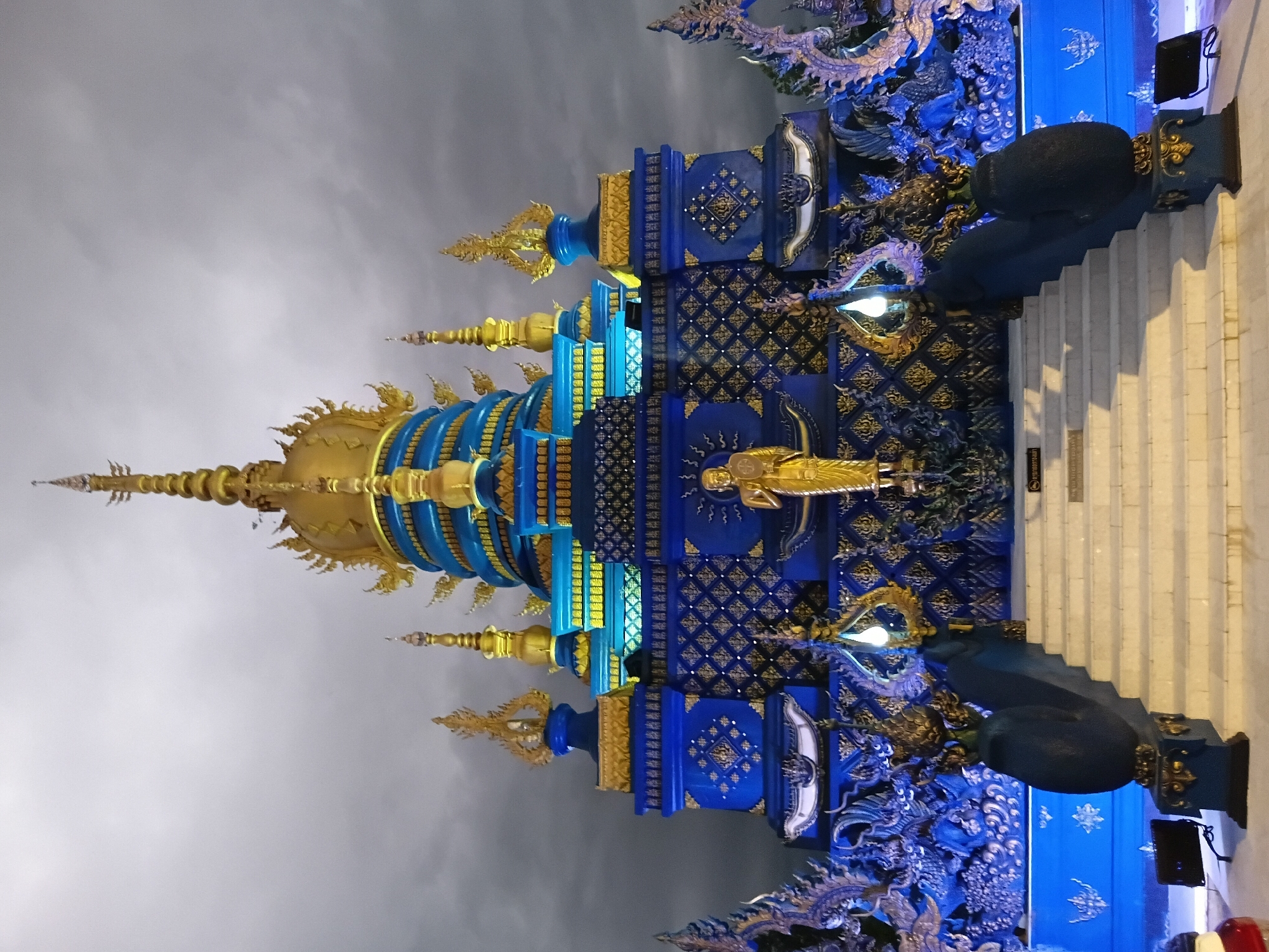 The image shows the Wat Rong Khun, also known as the White Temple, in Chiang Rai City Municipality, Thailand, at dusk. The temple is elaborately decorated with intricate blue and gold designs. A golden statue of a meditating monk stands prominently on a pedestal in front of the temple. The temple's tiered structure is topped with golden spires. The surrounding area is illuminated by blue and white lights, casting a vibrant glow on the temple's facades and reflecting on the wet pavement in the foreground. The sky is overcast, with dramatic clouds adding to the moody atmosphere. In the lower part of the image, a wide set of stairs leads up to the temple entrance, flanked by ornate blue sculptures that resemble mythical creatures. Several spotlights are positioned on the ground, illuminating the temple from below. The overall scene is one of striking architectural beauty and spiritual significance.