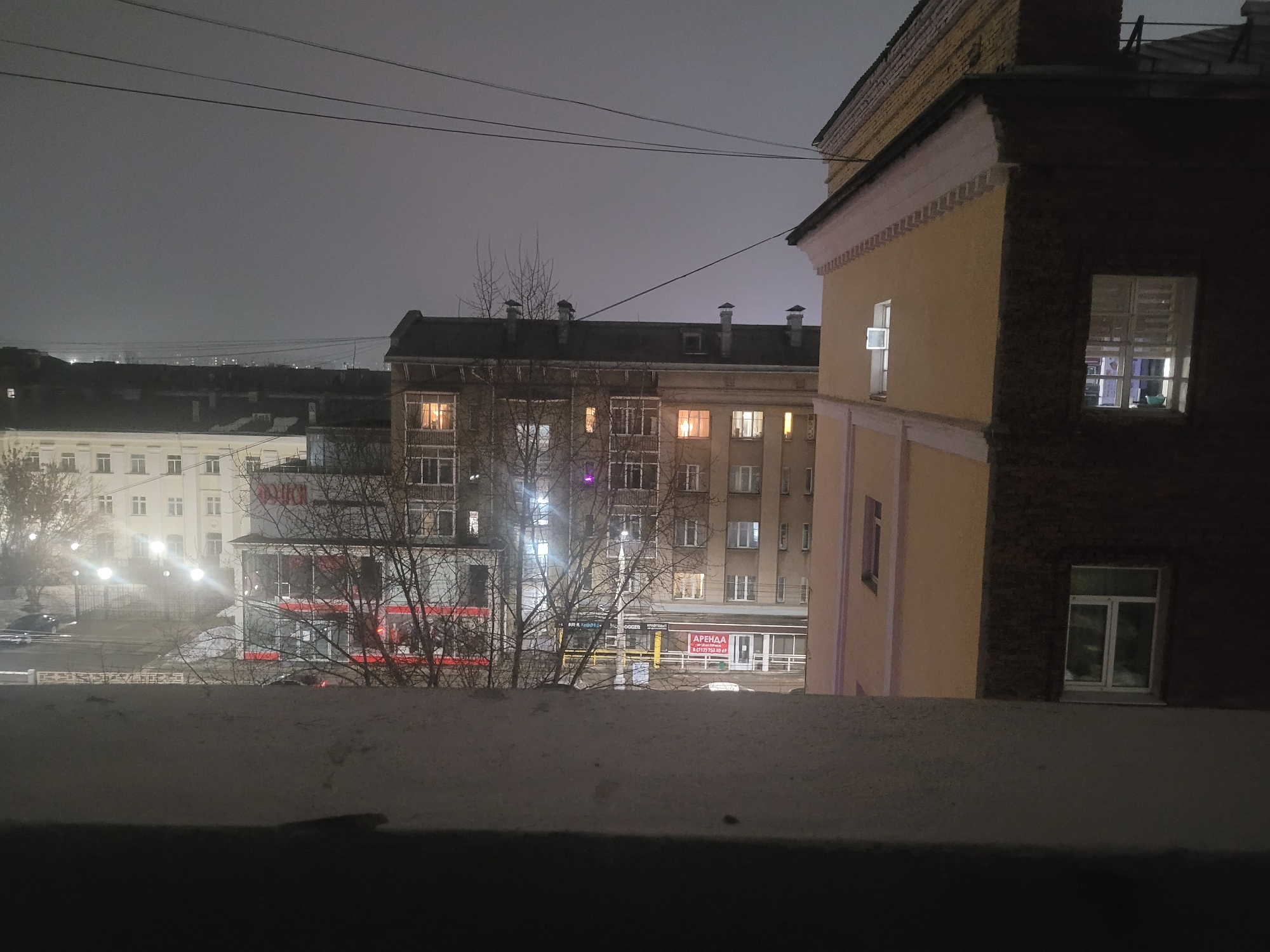 A nighttime cityscape in Izhevsk, Russia, viewed from an elevated position, possibly a window. The foreground shows a ledge, partially obscuring the view. In the middle ground are several multi-story buildings of varying colors (beige, grey, brown), some with lit windows. Bare trees are interspersed between the buildings. Street lights illuminate a street, and signage in Russian is visible on the lower levels of some buildings. The background is a dark, overcast sky.