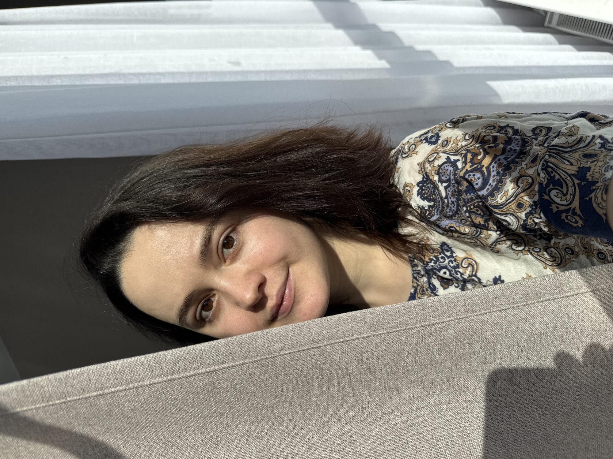 The scene portrays a female individual indoors in Izhevsk, Russia. The subject, an adult female with dark brown, shoulder-length hair and brown eyes, is positioned on the left side of the frame, partially visible as she peeks from behind a light beige, textured curtain. She wears a light-colored top with a blue, gold, and white paisley pattern. The subject looks directly forward with a slight smile. Strong natural light originates from the right side of the frame, illuminating the right side of her face and the curtain. On the right, a sheer white curtain covers a window, allowing sunlight to pass through and cast shadows. A white radiator or heating unit is visible at the bottom right corner of the frame.