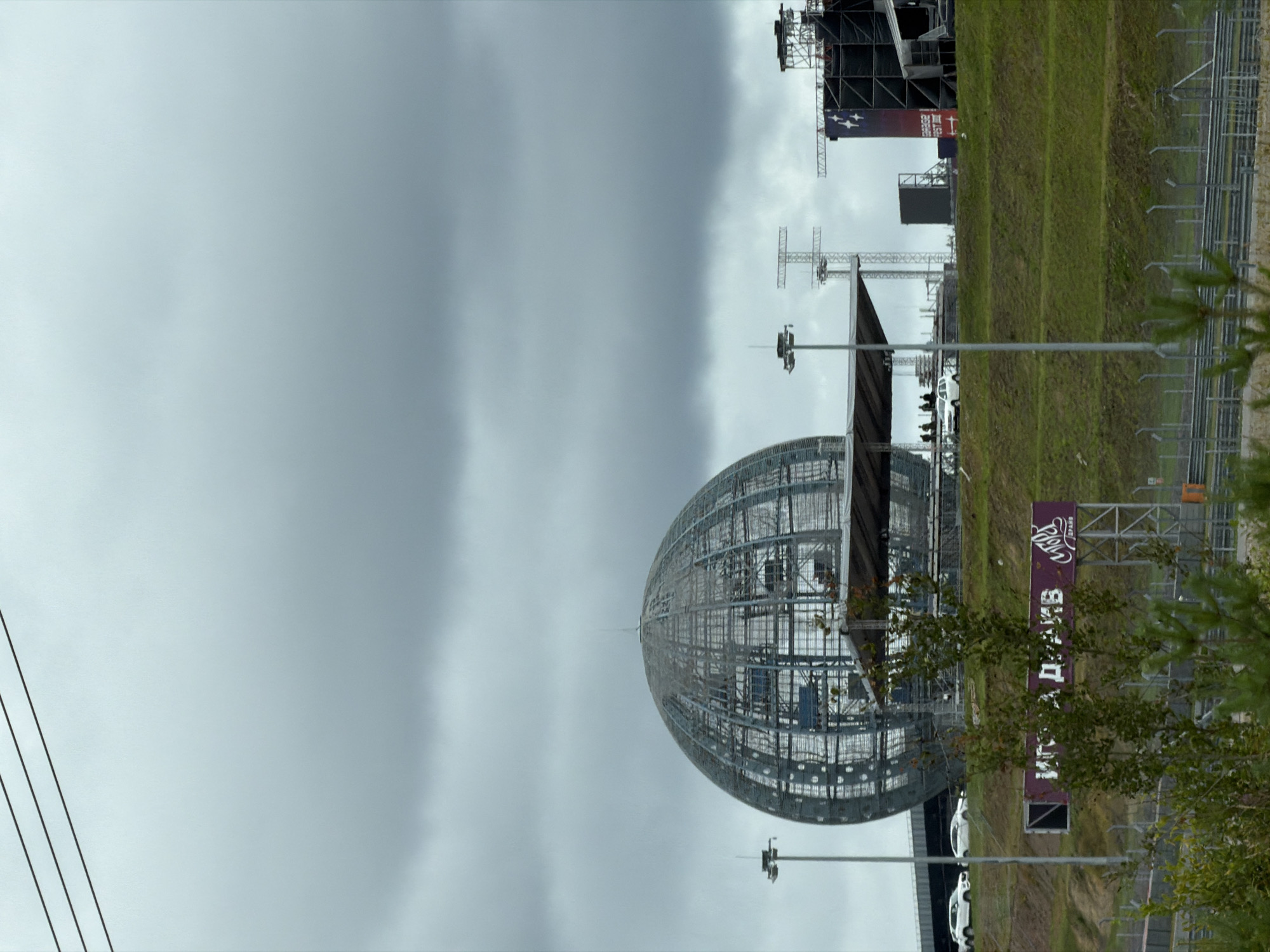 The image shows an outdoor scene under a cloudy sky, likely in Novozhilovo, Russia. In the background, a large, spherical, wireframe structure dominates the view. To the right of this sphere, a stage structure with lighting rigs and large screens is visible. In the mid-ground, a tiered grassy hill slopes upwards. Several white vehicles are parked on the crest of the hill, near a covered platform. People are standing on this platform. A sign with white text on a purple background is also present on the hill. In the foreground, there is a glimpse of a race track with fencing. Vegetation, including trees and grass, is scattered throughout the scene. Three dark, parallel lines are visible in the upper left corner of the image, possibly power lines.