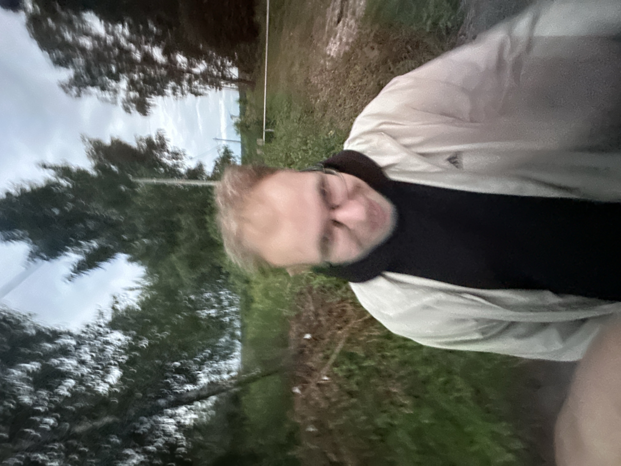 The image shows a person standing outdoors in what appears to be a rural or semi-rural setting, possibly near the city of Vostets Snt, Russia. The person is wearing glasses, a black hoodie, and a light-colored jacket. They are looking towards the camera with a slight smile. The background consists of trees, greenery, and a cloudy sky. The image is somewhat blurry, suggesting it was taken while moving or with a low-quality camera. There are no other people or distinct man-made structures visible in the immediate vicinity.