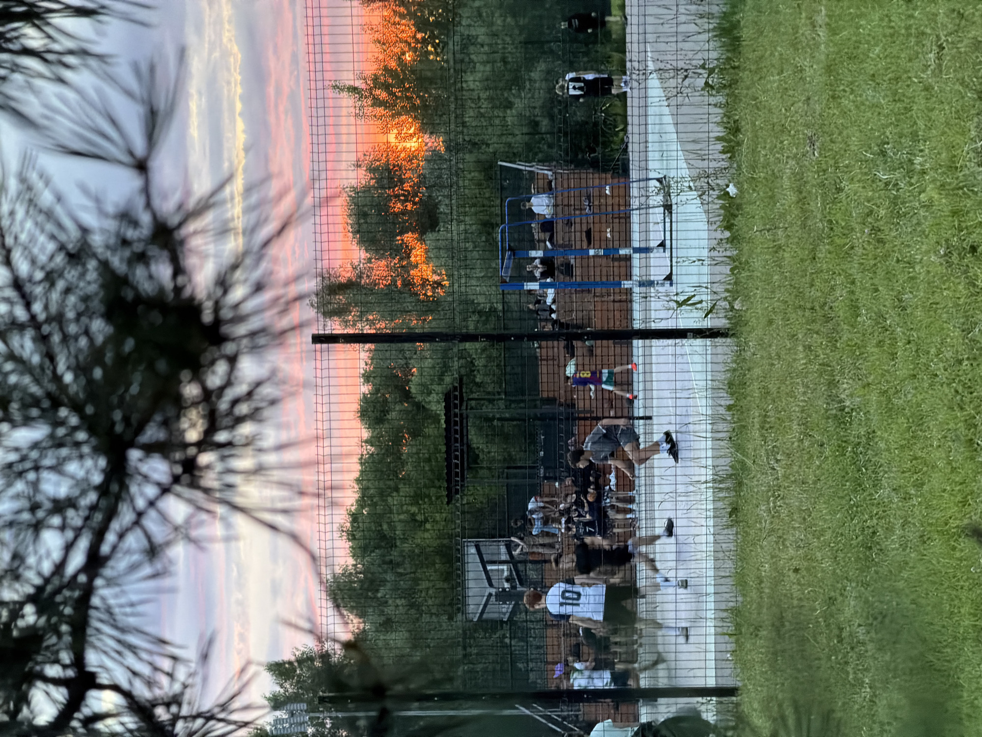 This media file shows an outdoor sports court with several people present. The scene is viewed through a wire fence, with a pine tree branch in the foreground, out of focus. The sky exhibits a sunset with vibrant orange and pink hues, suggesting it is evening.
The main subjects are a group of young people, appearing to be teenagers or young adults, engaged in some form of athletic activity. They are dressed in casual sportswear, including shorts and t-shirts, some with numbers on their jerseys. One person is wearing a white t-shirt with a large black "0" on the back. Another is wearing a purple and green jersey with the number "8". They are on a paved court that seems to be part of a larger sports complex, possibly a basketball or volleyball court, with some sort of net or wall in the background. There are also parallel bars, indicating a possible workout area.
The people appear to be actively participating in a game or practice. Their postures suggest movement and exertion. The overall mood seems to be energetic and social, typical of an evening sports gathering. The presence of trees and a grassy area surrounding the court points to a park or recreational facility. The location is specified as Kazan, Russia, and the sunset coloring of the sky is a prominent detail. There is no visible text that is clearly legible.