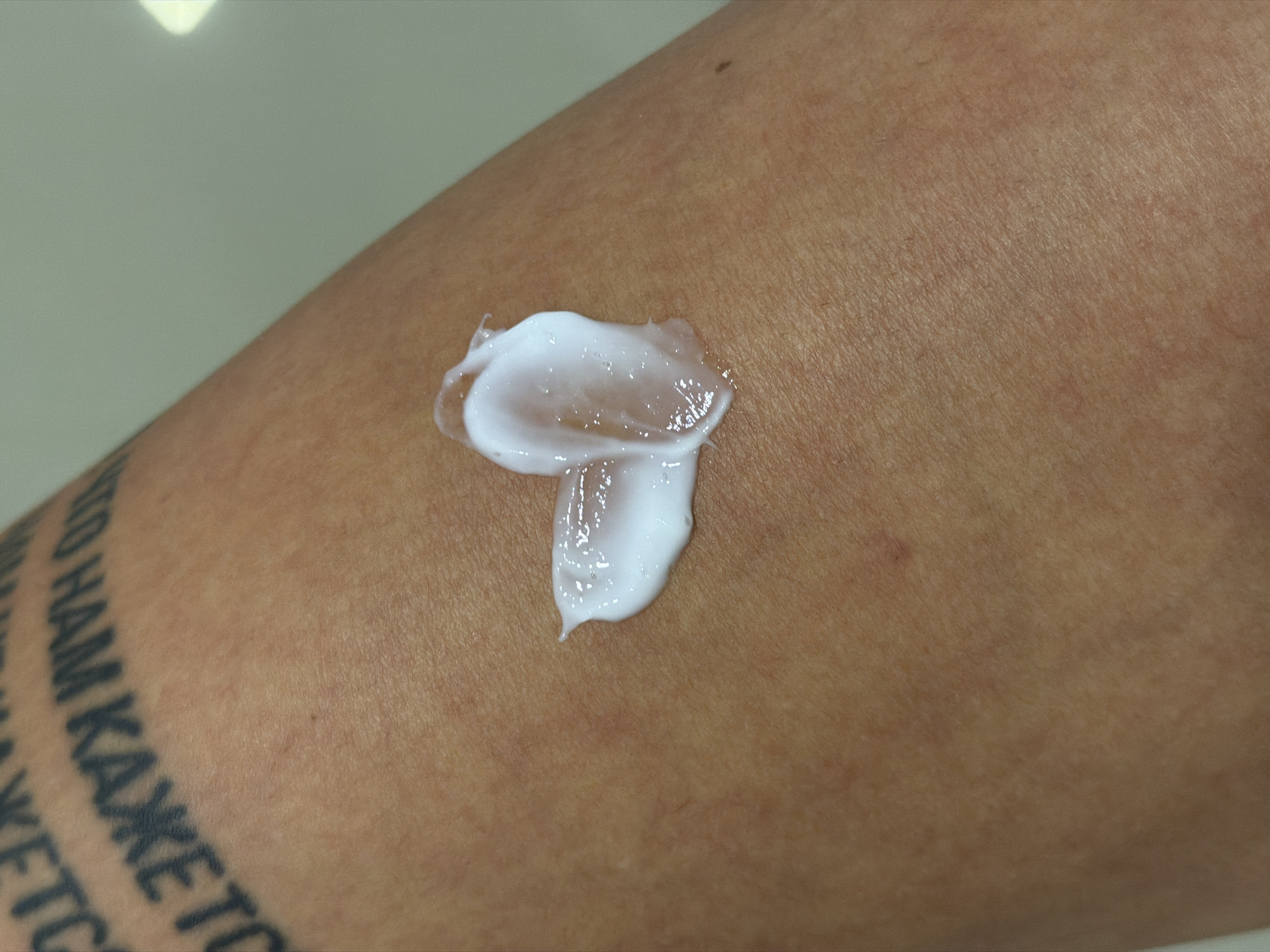 A close-up photograph depicts a light-skinned arm with a dab of white cream. The cream appears to be layered in two sections, the top layer is more opaque, and the bottom layer is thinner and more transparent. Part of a tattoo is visible on the lower left side of the image. The text of the tattoo, partially visible, is written in what appears to be the Cyrillic alphabet, and reads "КАЖЕТ". The skin shows pores and fine lines. The background is a blurred white. There are no indicators of location or that the scene is in Budva, Montenegro.