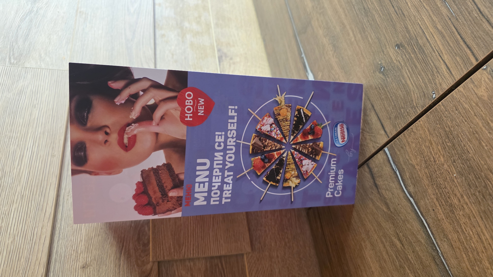 The image shows a promotional advertisement for Nestle Ice Cream cakes, specifically premium cakes, in the city of Burgas, Bulgaria. The advertisement features a woman with red lipstick and heavy makeup eating cake, next to the text "MENU" and "ПОЧЕРПИ СЕ! TREAT YOURSELF!". There is a stylized graphic of cake slices arranged in a circle, and a "NEW" badge in a heart shape. The advertisement is placed on a wooden surface, possibly a table or floor.