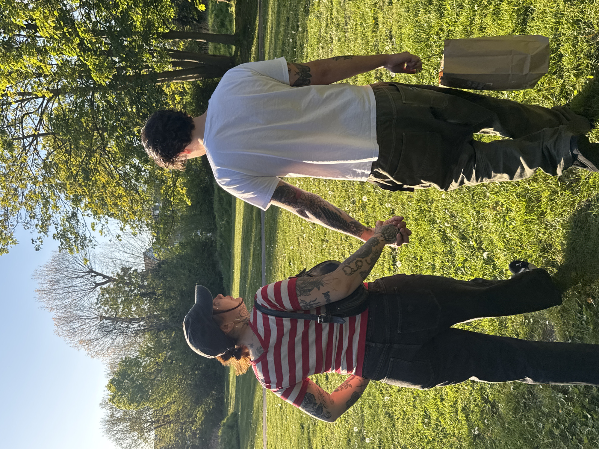 A couple is standing on a grassy lawn in a park under a bright blue sky. The person on the left is wearing a red and white striped shirt, black jeans, and a black baseball cap and has tattoos on their arms. The person on the right is wearing a white T-shirt and green pants, also with tattoos on their arms. They are holding hands. Next to the person in the white T-shirt is a brown paper bag. The park has trees and dandelions. The location appears to be a public park in London, United Kingdom.