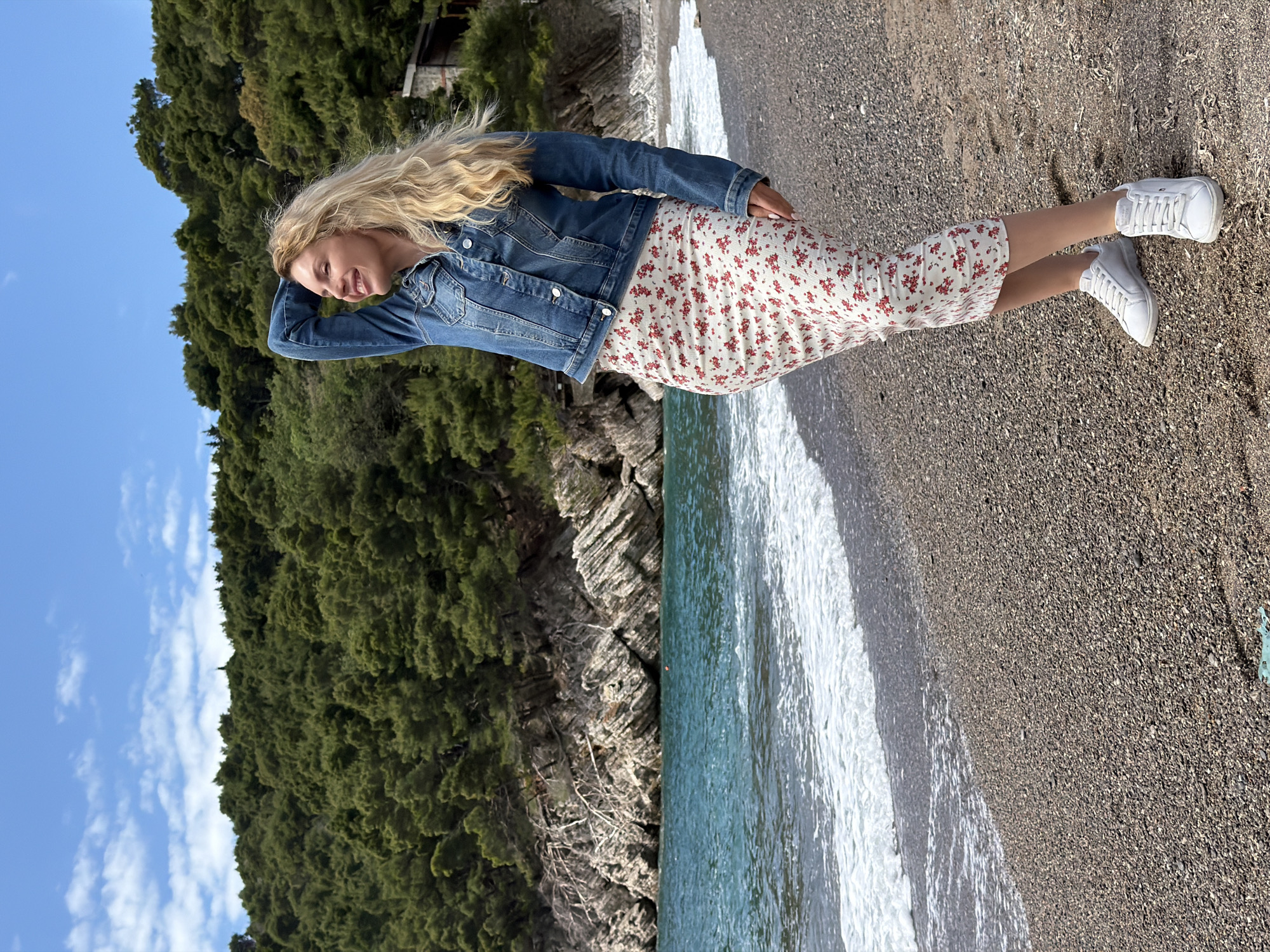 A woman stands on a beach in Petrovac centar, Montenegro. She is wearing a denim jacket, a white floral dress, and white sneakers. Her blonde hair is long and wavy. She is looking to the side, with her hand on her forehead, seemingly shading her eyes. The beach is sandy with small pebbles. Waves are lapping on the shore. The water is turquoise. Behind her is a rocky cliff covered with lush, green vegetation. The sky is blue with some scattered clouds.