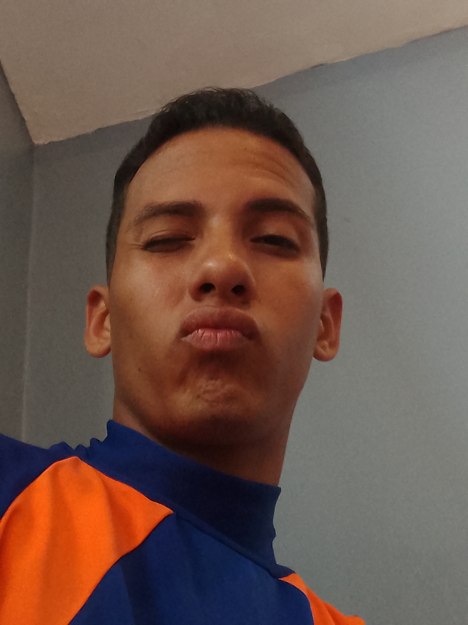 A close-up selfie depicts a man with brown skin, dark hair, and a pursed lip expression. He is wearing a blue and orange athletic-style shirt. The background consists of a gray wall and a partially visible white ceiling. The exact location within the city of Porlamar, Venezuela cannot be determined from this image alone.