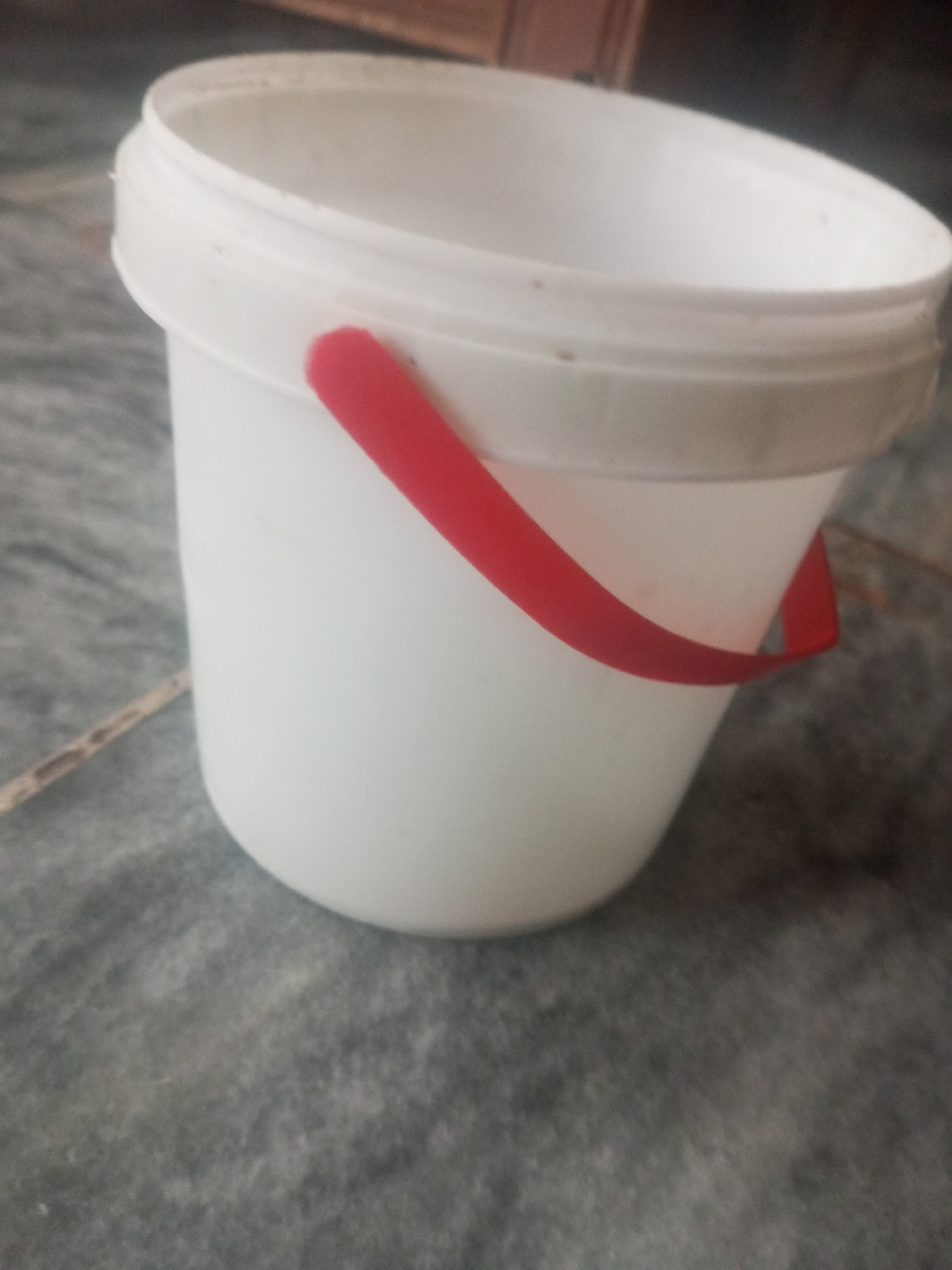 A close-up, slightly blurry, eye-level shot shows a white plastic bucket with a red handle. The bucket is sitting on a gray, textured surface that appears to be a tiled floor. The background is dark and out of focus, but suggests an indoor environment, possibly a room. There are no people or other objects clearly visible in the scene. The image does not provide any specific information about the location in Mansehra, Pakistan.