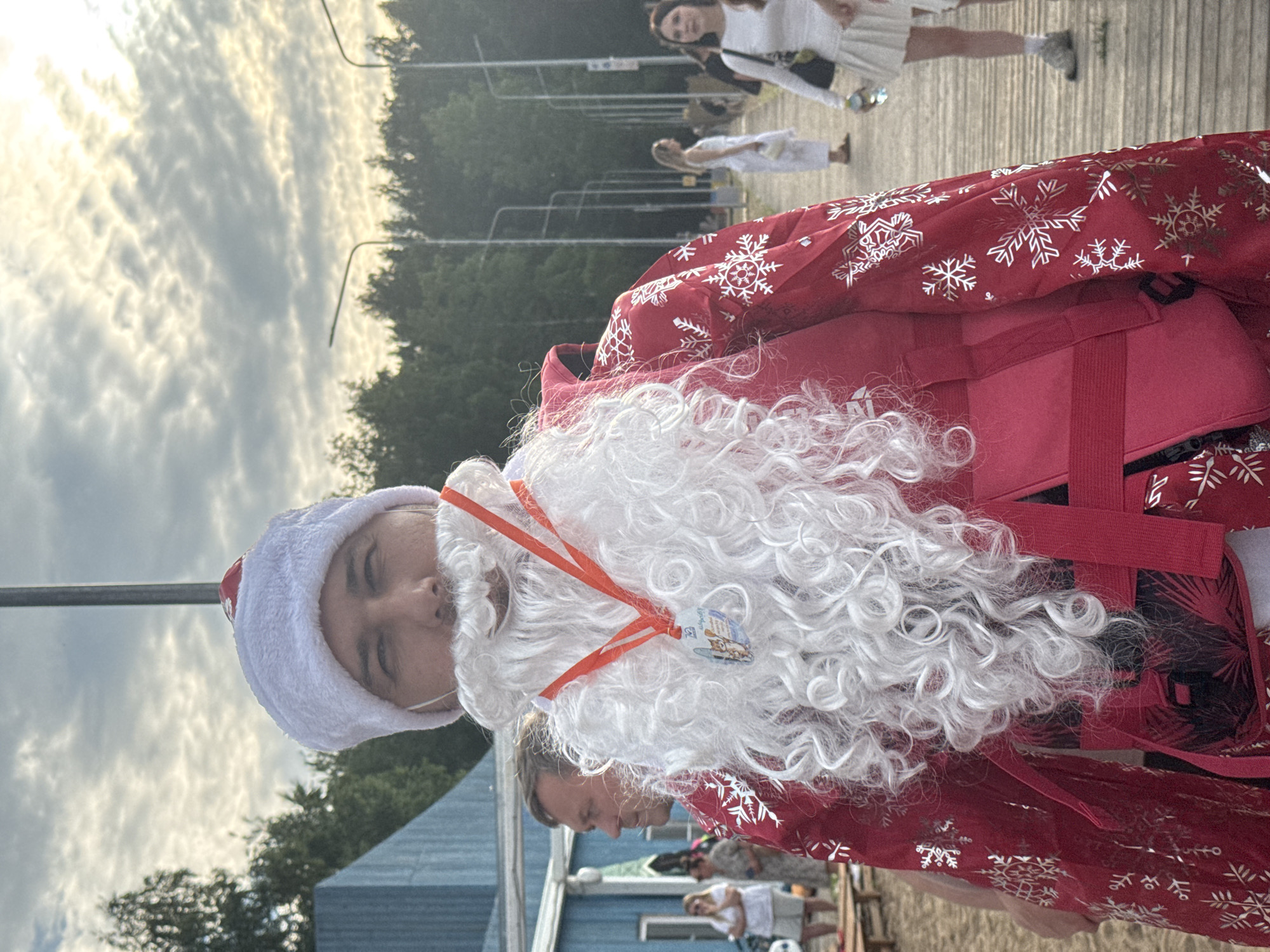In Chaikovsky, Russia, a man in a Santa Claus costume stands on a wooden boardwalk under a cloudy sky. He wears a white fur-lined hat and a large, curly white beard. A red jacket adorned with white snowflakes covers his upper body, cinched with a red sash. A medal on an orange ribbon hangs around his neck, partially obscured by his beard. In the background, a blue building is visible on the left, and trees line the right side of the boardwalk, where several people are walking. The setting appears to be an outdoor public space, possibly near a beach or a waterfront.
