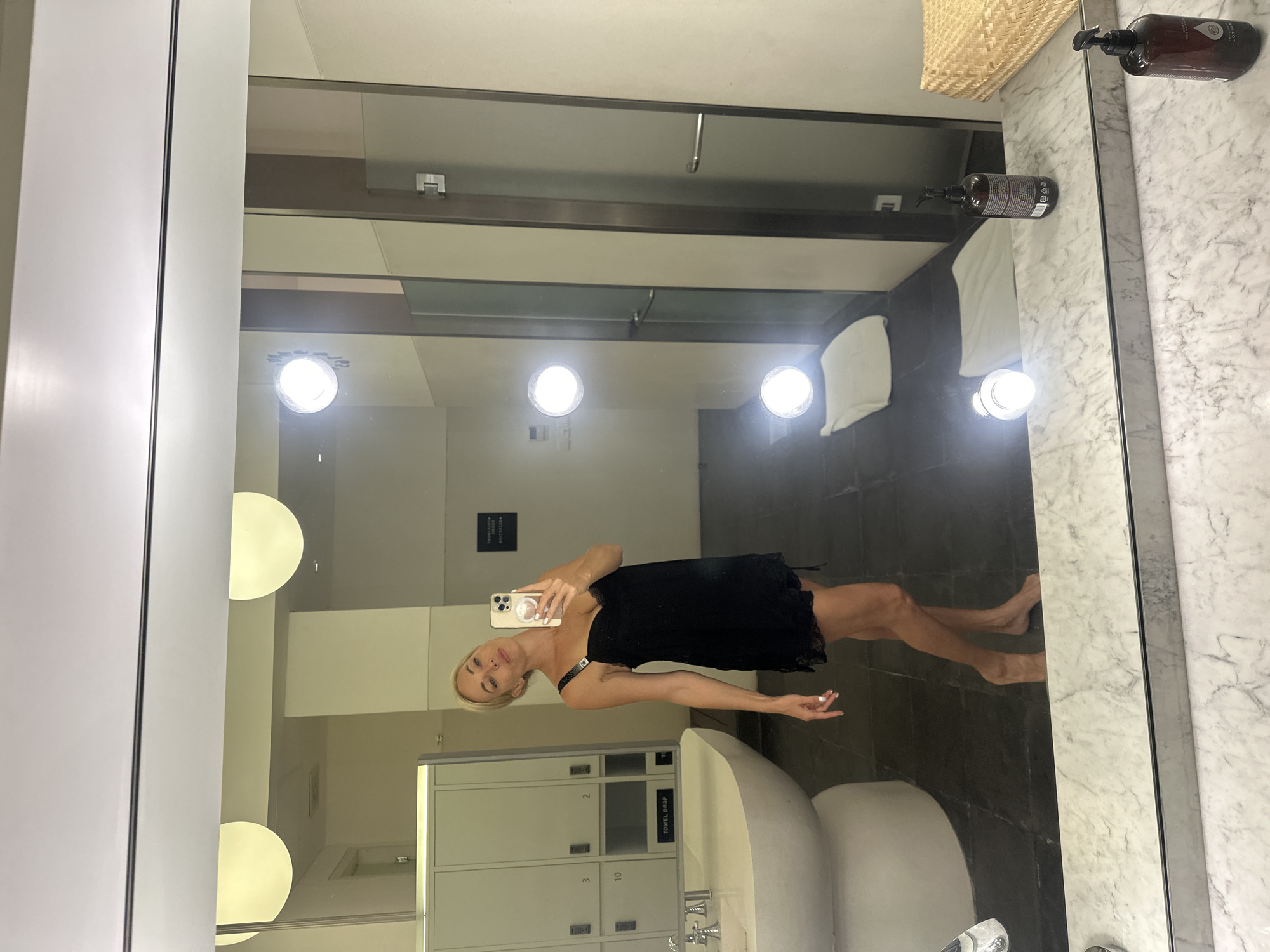 A woman is taking a selfie in a locker room with a large mirror. She is wearing a black slip dress and has blonde hair. The locker room has lockers, a sink, and bright lights. The mirror reflects the woman and the locker room. The location cues suggest it is a spa or gym locker room. The time of day is not discernible. The woman's expression is neutral, and she is posing for the photo. There is a brown bottle with a pump dispenser on the counter next to the mirror. The text "TOWEL DROP" is visible on one of the lockers.