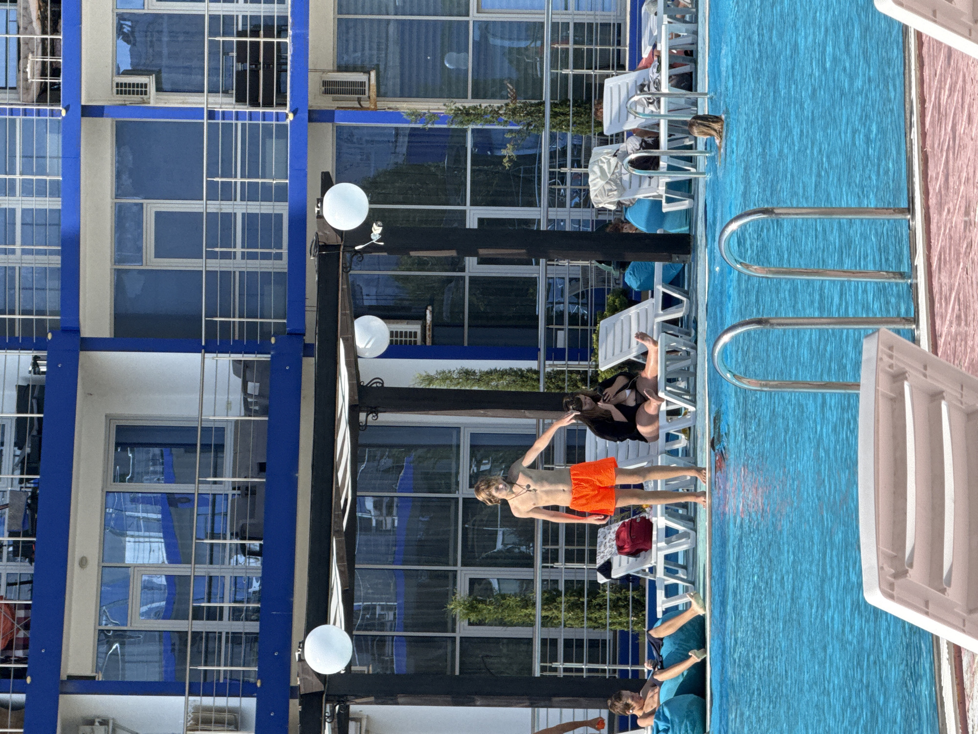 The image depicts a sunny outdoor scene at a hotel or resort with a swimming pool. In the foreground, a bright blue swimming pool with a pink tiled edge is visible, featuring metal pool ladders. Several white lounge chairs are positioned around the pool, some occupied by people.
In the middle ground, a young man with blond hair, wearing bright orange swim trunks, stands near the edge of the pool, facing to his right and gesturing with his left hand. Behind him, a woman in a black cover-up is seated on a lounge chair, looking at her phone. To the left, a young boy is reclining on a blue beanbag chair.
The background shows a multi-story building with blue accents and balconies, common in resort architecture. The building has numerous windows and several air conditioning units. The scene appears to be set in a warm climate, with clear skies and bright sunlight casting shadows. The overall impression is of a relaxed holiday environment. The location is identified as Sevastopol, Ukraine.