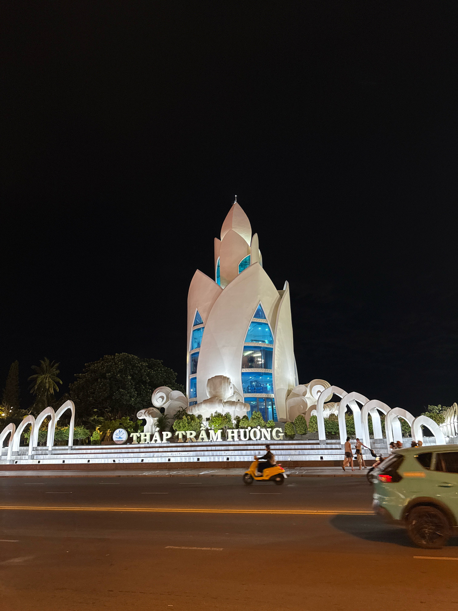 The image shows the Po Nagar Cham Towers, a historical complex located in Nha Trang, Vietnam, at night. The central tower, illuminated and architecturally distinctive with its petal-like structure and blue glass windows, is the focal point. In front of the towers, a large sign reads "THAP TRAM HUONG" in illuminated letters, with decorative arches flanking it. The scene includes a road in the foreground with a yellow scooter carrying a rider, and a light-colored SUV driving by. Several people are visible walking near the sign and arches. Lush green trees and palm trees are present in the background, and the sky is dark, indicating nighttime.
