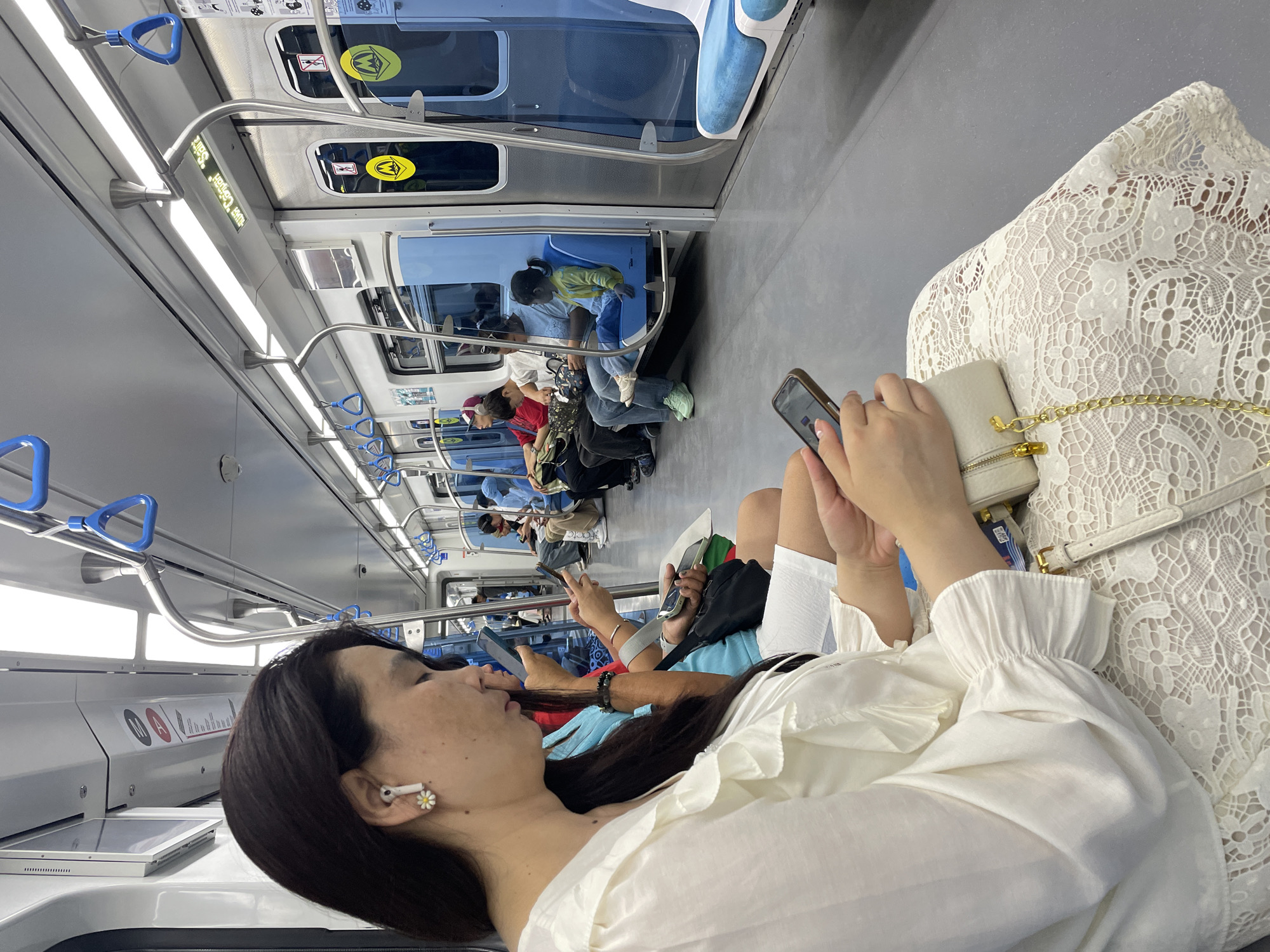 An interior view of a modern, clean subway train carriage in Almaty, Kazakhstan, shows passengers seated. The main subject is a woman in the foreground, wearing a white blouse with ruffled sleeves and a white lace skirt, looking down at her smartphone. She has dark hair and is wearing a white earbud with a small daisy charm. Beside her, another woman in a blue top is also looking at her phone. Further in the carriage, various other passengers are visible, including a child in a yellow jacket sitting on a blue seat. The train carriage features metallic grab handles, overhead lighting, and advertising displays above the doors. The flooring is a light grey material, and the seats are a combination of blue and grey. The overall impression is one of orderly public transport.