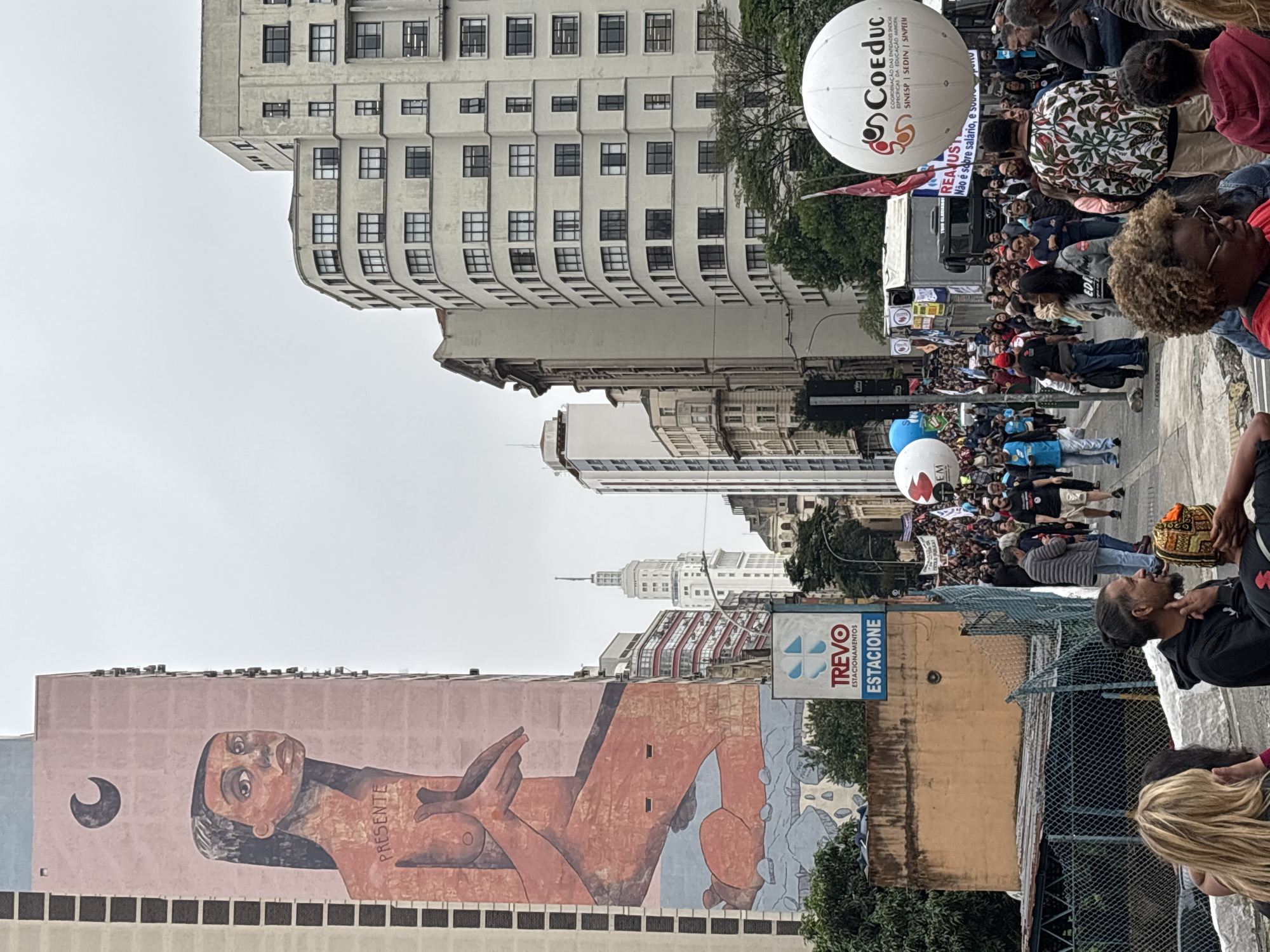 The image captures a street scene in São Paulo, Brazil, marked by a protest or march. A large crowd of people fills the street, with two large white balloons bearing the word "Coeduc" visible among them. The street is lined with tall buildings, one with a mural of a reclining figure. A sign for "Trevo Estacione" (Trevo Parking) is visible. The sky is overcast.