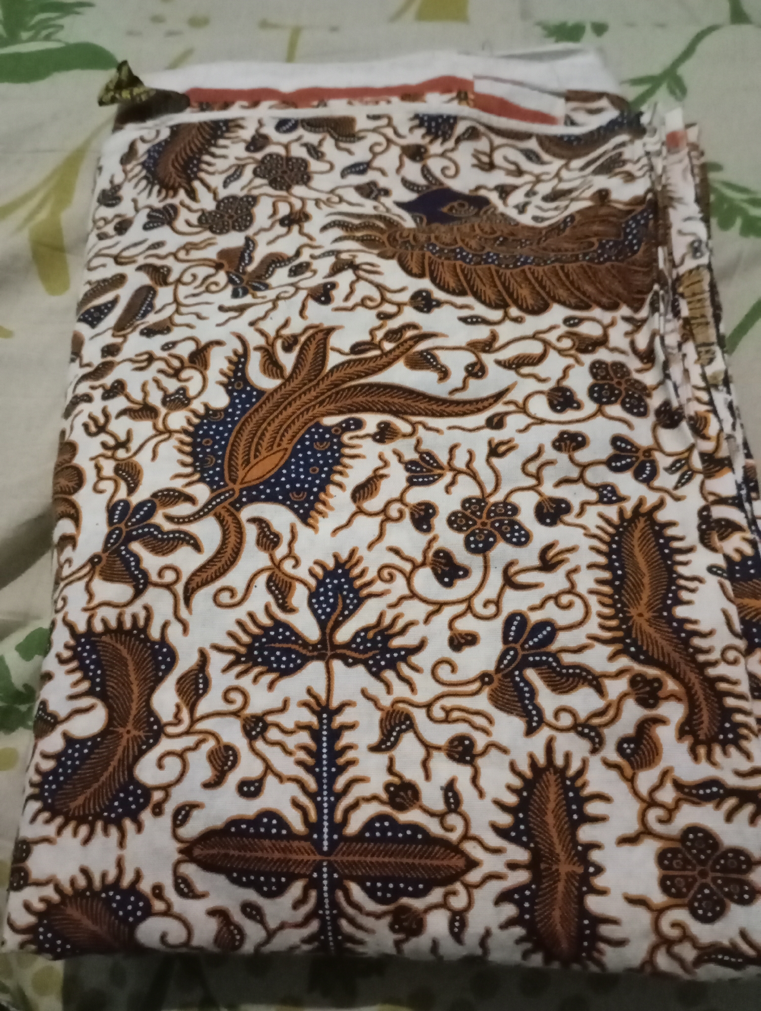 The image shows a folded piece of white fabric with a traditional batik pattern. The pattern features brown and dark blue stylized motifs resembling birds and floral elements, set against the white background. The fabric is neatly folded, with its edges visible. A small, folded piece of fabric, possibly part of another textile, is visible at the top left corner of the image, showing a green and yellow pattern. The background fabric beneath the folded batik appears to be a light-colored textile with a faint green leaf-like pattern. The scene is indoors, and the lighting suggests it could be natural daylight or artificial light. There are no people present in the image. The context of Kecamatan Haurgeulis, Indonesia, is not visually discernible from the image itself, as it only displays the fabric.