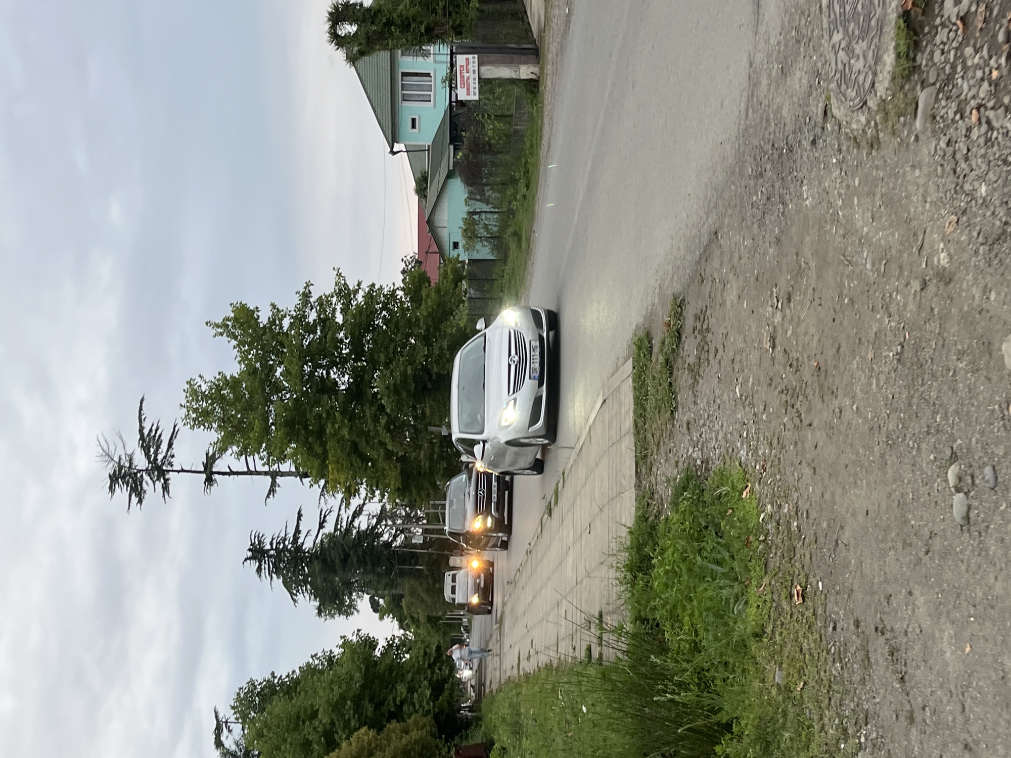 The image depicts a street scene in Ureki, Georgia. A silver Opel Insignia is driving on the right side of the road. A black Mercedes-Benz SUV is in front of it, followed by a white van and a yellowish vehicle. A person is walking on the sidewalk to the left of the road, which is lined with trees and grass. In the background, there are houses and more trees under a cloudy sky. A sign with text is visible on the fence of one of the houses. The road surface is asphalt, and the sidewalk is paved with rectangular stones.
