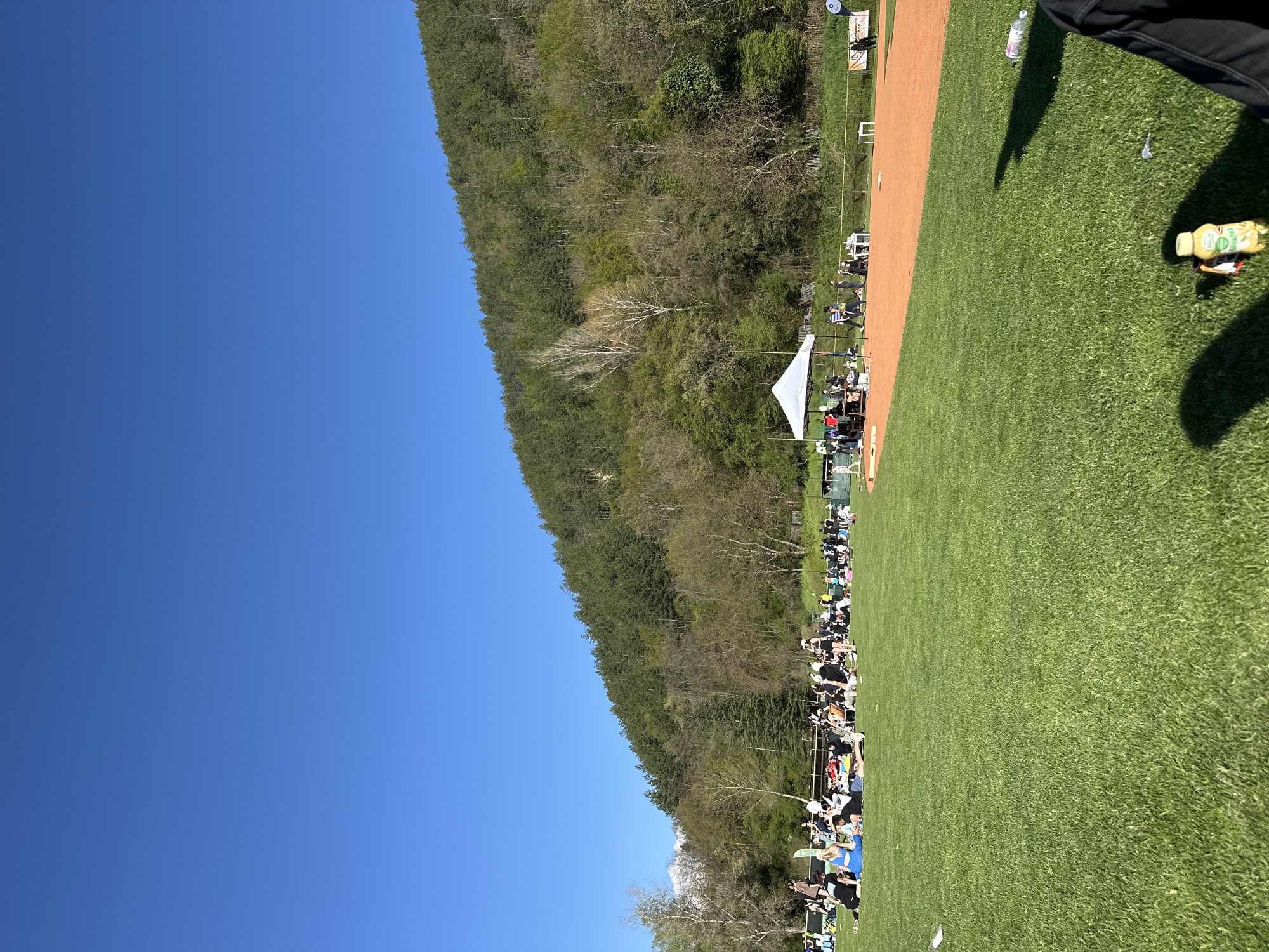 The image shows an outdoor baseball game in Blagoevgrad, Bulgaria. A green field stretches to the right, adjacent to a red-clay infield with a baseball diamond visible. People are seated along the edge of the field, and a forest rises in the background against a clear blue sky. A white tent stands near the crowd.