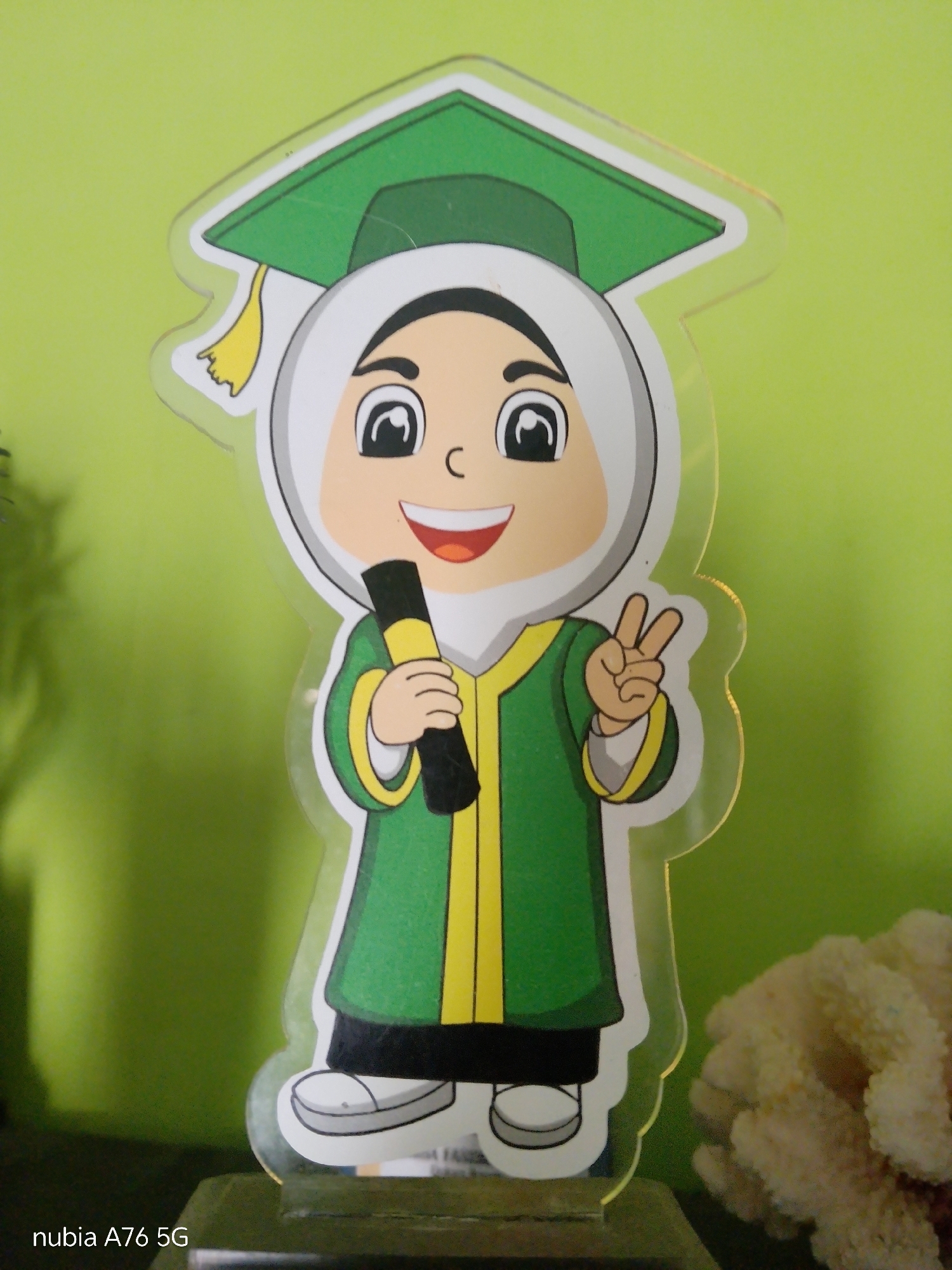 The image is a close-up, eye-level, vertical shot of a graduation-themed acrylic standee featuring a cartoon character. The character is a young girl wearing a green graduation cap with a yellow tassel and a white hijab. She is also dressed in a green graduation robe with yellow trim and is holding a black rolled-up diploma with a yellow band. Her left hand is making a peace sign. The standee is placed on a dark brown base, which is slightly visible at the bottom of the image. The background is a bright, solid green wall. In the lower right corner of the image, a portion of a textured, off-white object, possibly a decorative coral or sponge, is visible. The text "nubia A76 5G" is subtly imprinted in the bottom left corner of the image, indicating the device used for capture. This image was captured in the city of Cakung, Indonesia.