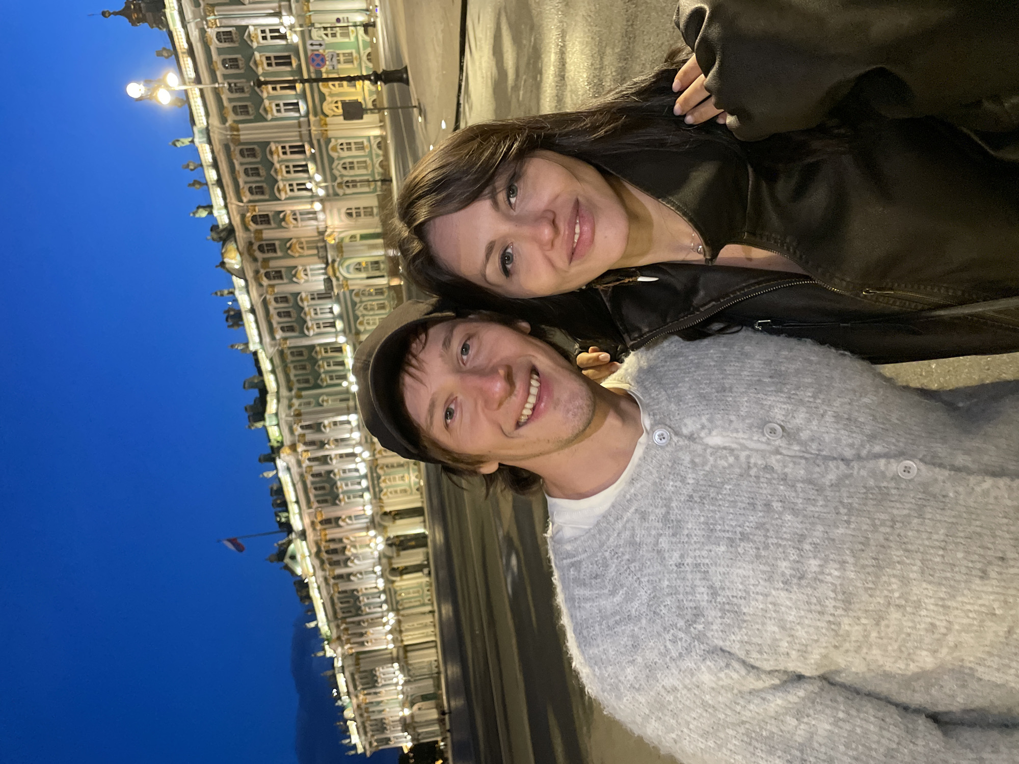 A couple is taking a selfie in front of the Winter Palace at dusk. The man, on the left, is wearing a grey cardigan and a black baseball cap. The woman, on the right, is wearing a black leather jacket. The Winter Palace, a former imperial residence, is illuminated and stands majestically in the background. The sky is a deep blue, transitioning from daylight to night. The Palace Square, a prominent public space in Saint Petersburg, Russia, is visible. The ground is wet, suggesting recent rain or reflection from lighting.