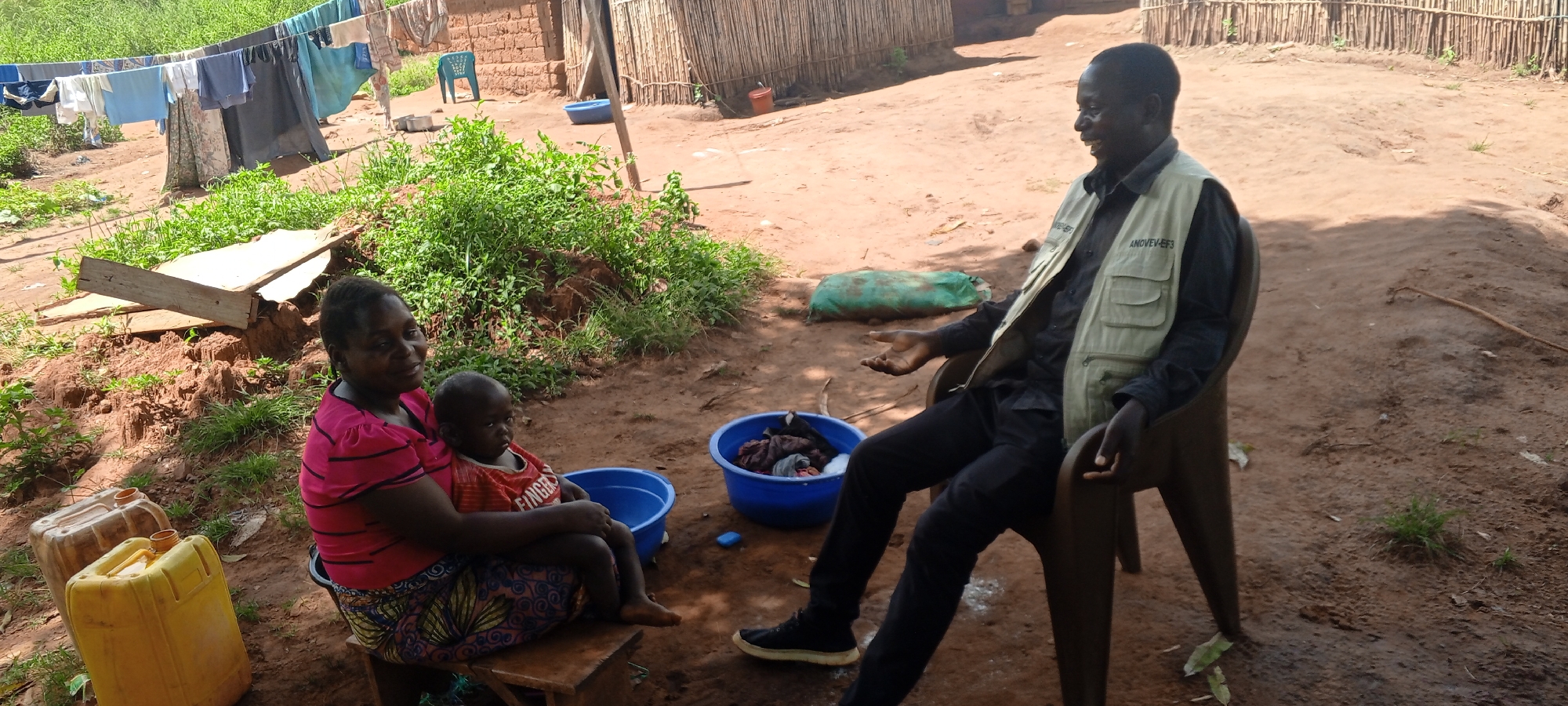 The image shows a man, woman, and child in what appears to be an outdoor setting in the city of Makungu, Democratic Republic of the Congo. The man is sitting on a chair and wearing a vest with the text "ANOVEVEF3" on the left breast. The woman is seated on a wooden stool and holding the child. Next to them are two yellow jerrycans and a blue washbasin. A clothesline with hanging garments is visible in the background.