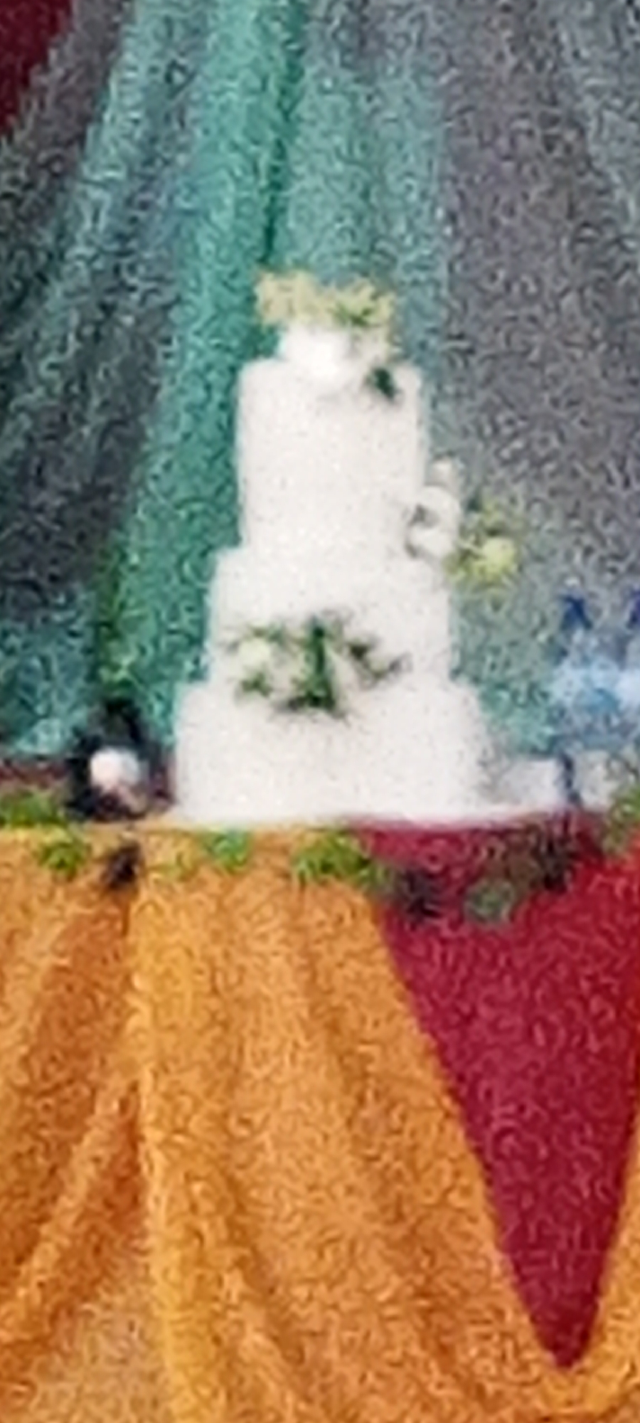 A blurry image of a three-tiered white cake decorated with flowers. The cake sits on a table covered with orange and red fabric. The background is a curtain, possibly teal or blue. The precise location and details of Kakuri Gwari, Nigeria, are not discernible from the image. No people are visible.