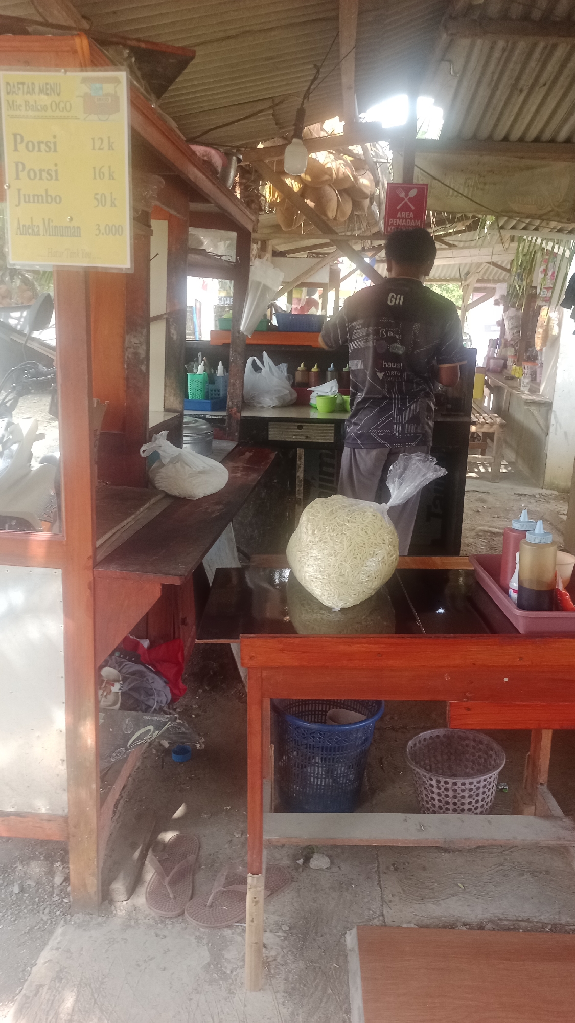 In the city of Cibeber, Indonesia, a street food stall selling mie bakso, or meatballs noodles, is depicted. The stall has a rustic appearance, constructed from wood with a corrugated metal roof. A yellow sign listing menu items and prices for "Mie Bakso OGO" is prominently displayed on the left side. The menu items include "Porsi" priced at 12k and 16k, "Jumbo" at 50k, and "Aneka Minuman" (various drinks) at 3,000.
In the background, a person wearing a dark t-shirt with "GII" on the back is preparing food. They stand behind a counter laden with food preparation items. To the right of the counter, a sign with a red symbol indicates "AREA PEMADAM KEBAKARAN" (Fire Extinguisher Area). On a wooden table in the foreground, a large clear plastic bag filled with raw noodles is placed.
Various other items are scattered around the stall, including white plastic bags, bottles of sauces, plastic baskets, and a pair of flip-flops on the ground. The overall environment suggests a typical outdoor street food setting.