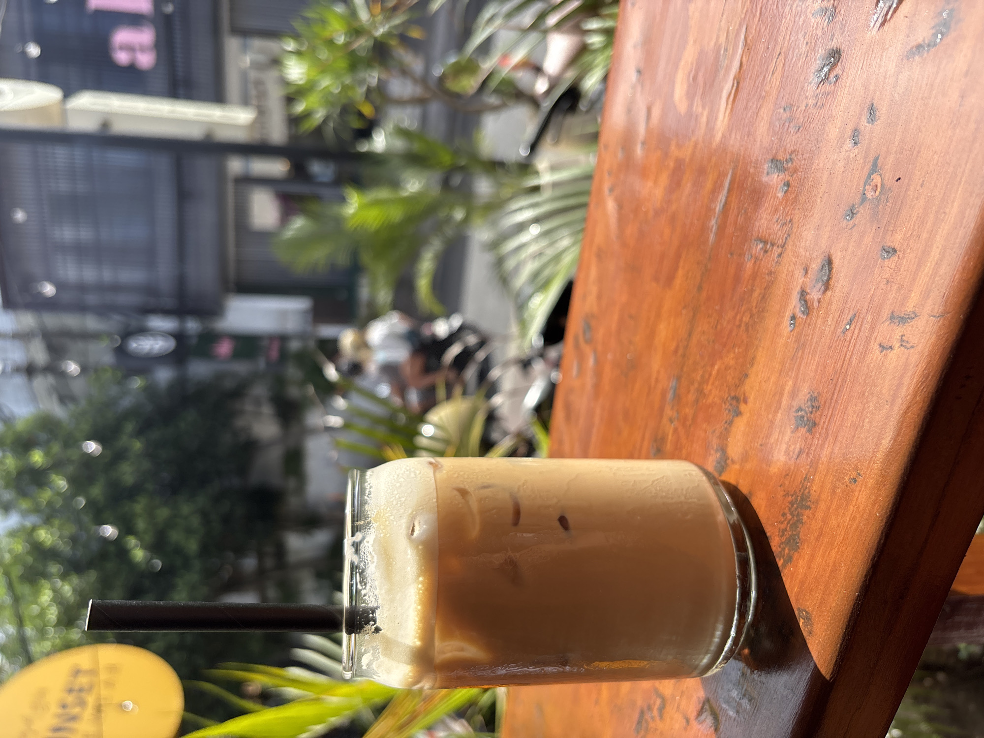 A glass of iced coffee sits on a weathered, varnished wooden surface. A black straw is visible in the drink. The background shows an outdoor scene with greenery and blurred buildings, suggesting an urban setting. The photograph seems to have been taken in daylight, with strong sunlight illuminating the wooden surface. The location is likely Kuta Selatan, Indonesia. There are no people distinctly visible, but there are people blurred in the background.