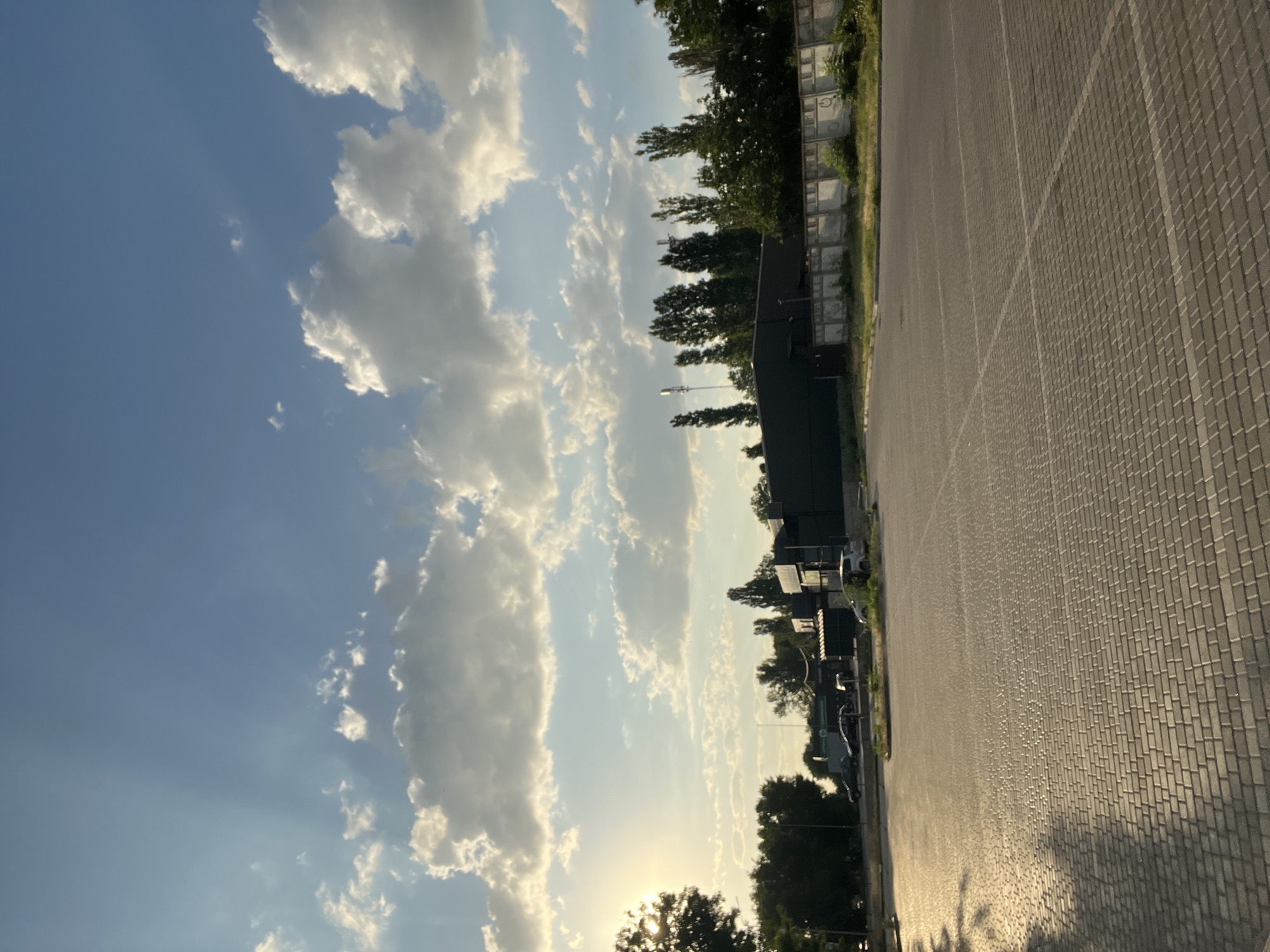 This is a ground-level, eye-level, full shot image of a paved outdoor area, likely a parking lot, in Fontanka, Ukraine. The sky above is a light blue with scattered white cumulus clouds, some of which are illuminated by the sun, creating crepuscular rays that extend downwards. The sun is visible in the upper left quadrant, partially obscured by a tree's foliage, casting a warm, golden light across the scene and creating long shadows.
In the mid-ground, there is a dark, rectangular building with a light-colored fence or wall in front of it. The fence has a weathered appearance and appears to be made of concrete blocks. Behind the fence and to the right of the building, there is a line of tall, slender trees. Several cars are parked in the area in front of the building, with one white car positioned closer to the foreground.
The foreground is dominated by a gray, interlocking paver surface, arranged in a repeating pattern. Faint white lines marking parking spaces are visible on the pavement. The overall atmosphere is calm and still, suggesting it is either early morning or late afternoon due to the quality of the sunlight and the length of the shadows. There are no people visible in the image.