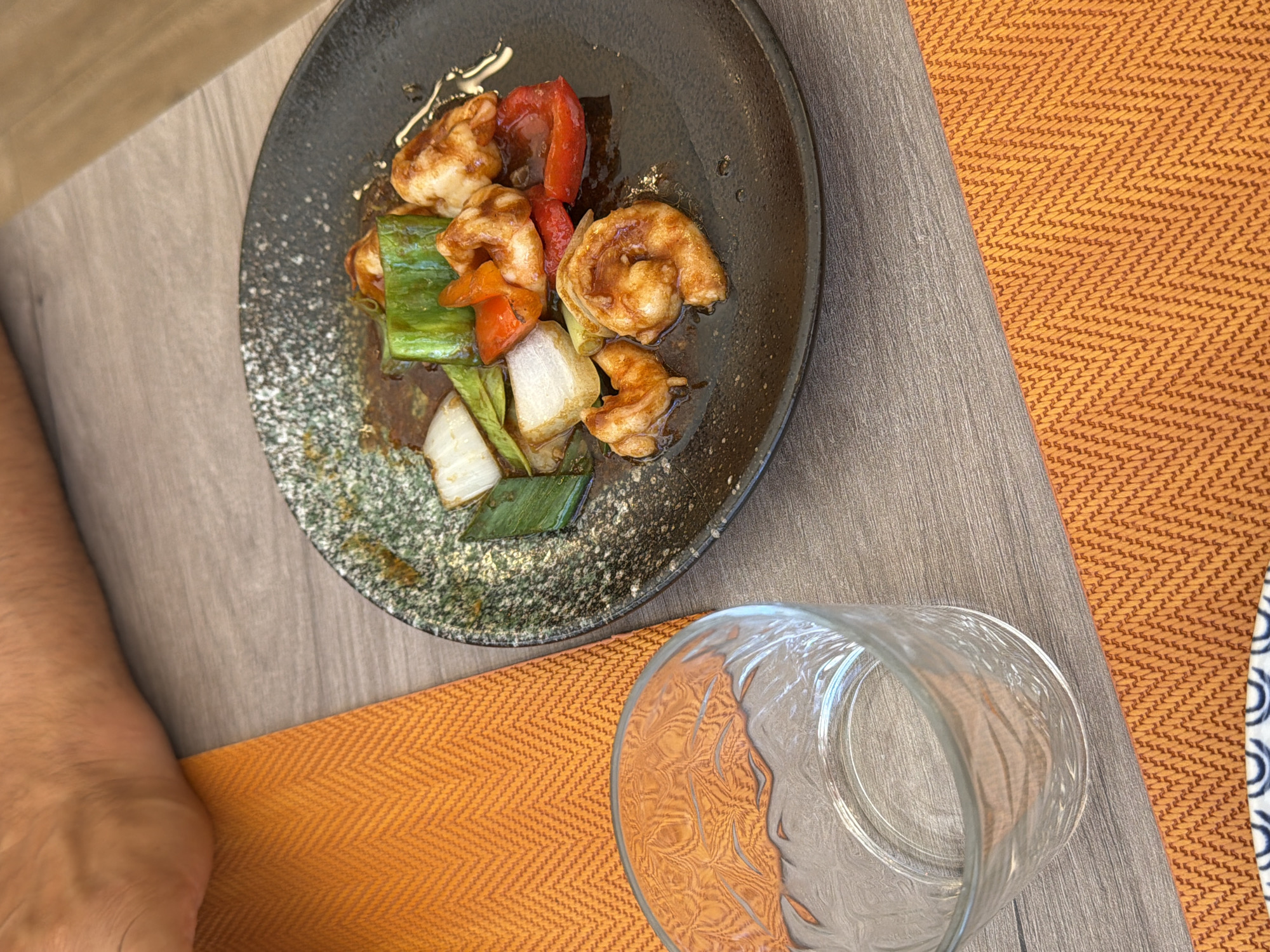 A close-up, overhead shot shows a plate of stir-fried shrimp with vegetables, resting on a dark, speckled plate. The dish contains several pieces of shrimp, chunks of red bell pepper, white onion, and green bell pepper, all coated in a glossy sauce. The plate is positioned on a grey wooden surface. To the left of the plate, an orange, textured placemat is visible, and a clear glass with an embossed pattern sits on top of it. In the background, a person's forearm and hand are partially visible, resting near the placemat. The scene appears to be set in an outdoor dining area, likely at a restaurant in Benidorm, Spain, suggested by the bright, warm lighting and the presence of a woven placemat often found in coastal Mediterranean establishments.