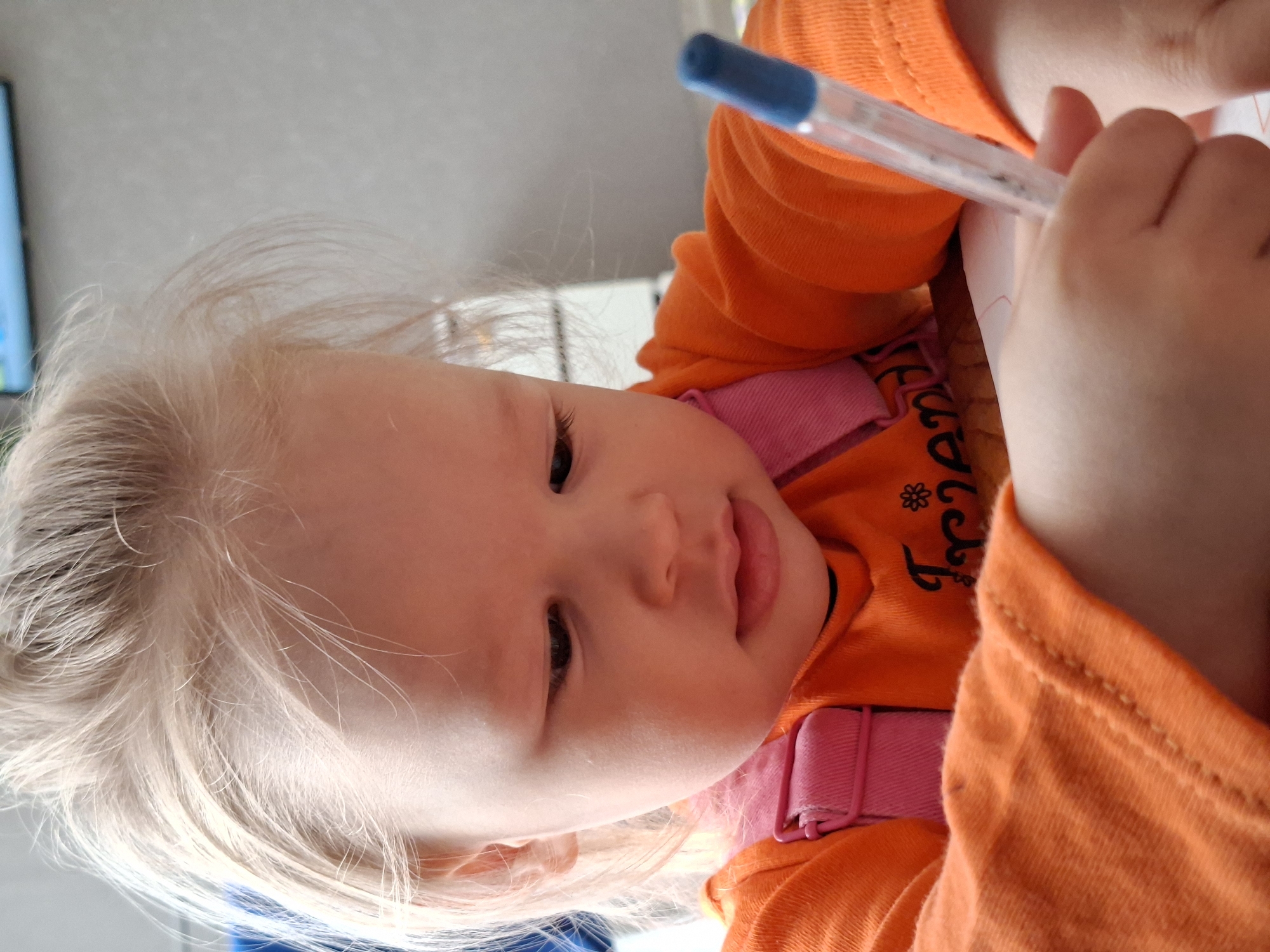 A close-up shot depicts a toddler with fair skin and blonde hair, presumably drawing or writing with a pen. The child is wearing an orange shirt with a pink harness or straps visible. The setting is not specified or determinable from the image.