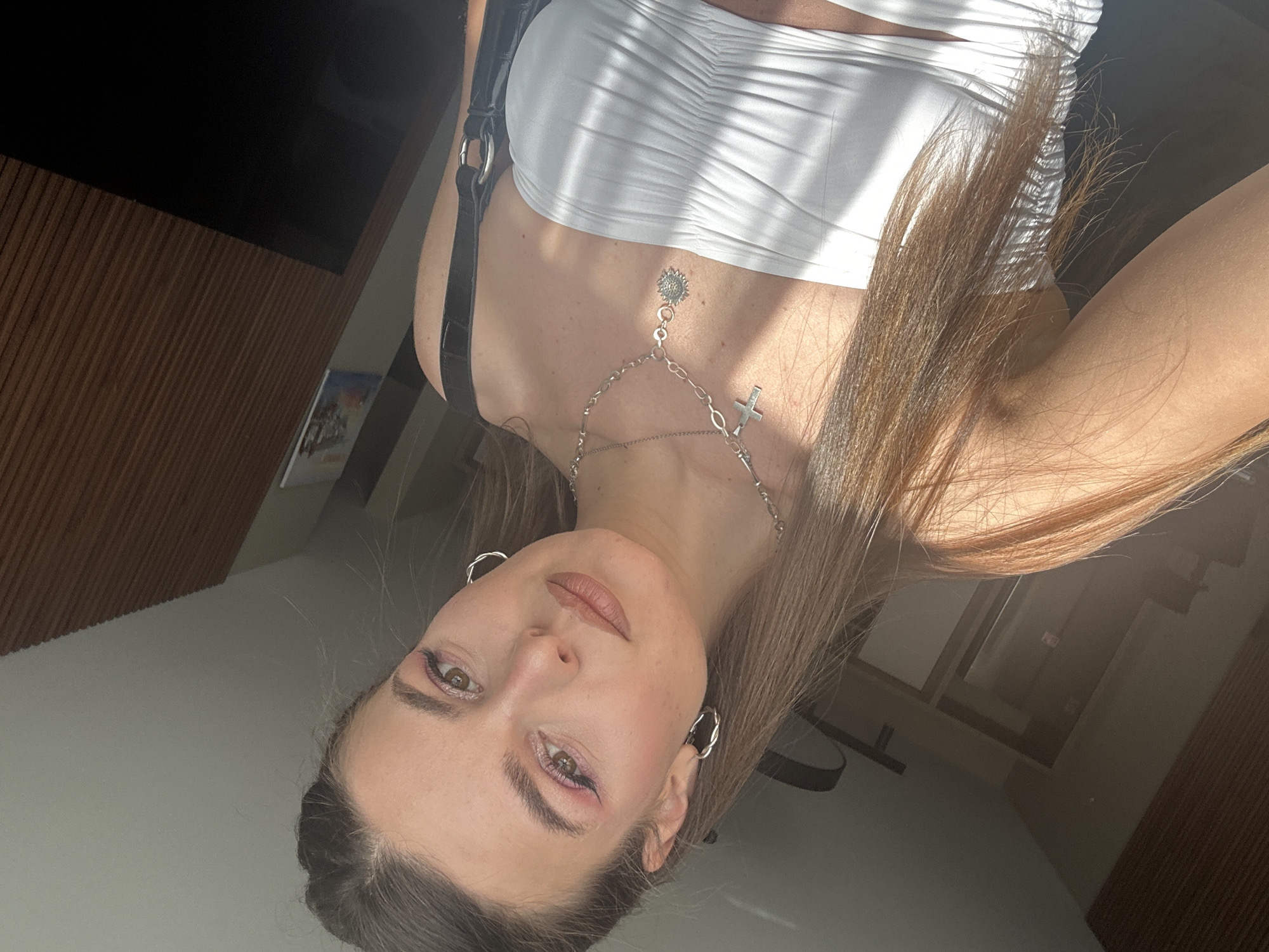 A young woman is facing the camera while lying down, with her head tilted to the left. She has light brown hair, fair skin, and is wearing silver hoop earrings and a silver necklace with a cross pendant. She has a small, circular tattoo at the base of her neck. She is wearing a white, ruched top with a black strap visible. The background appears to be an indoor setting with wooden paneling and a glimpse of a window. Without additional information, it is impossible to determine whether this photo was taken in Plovdiv, Bulgaria.