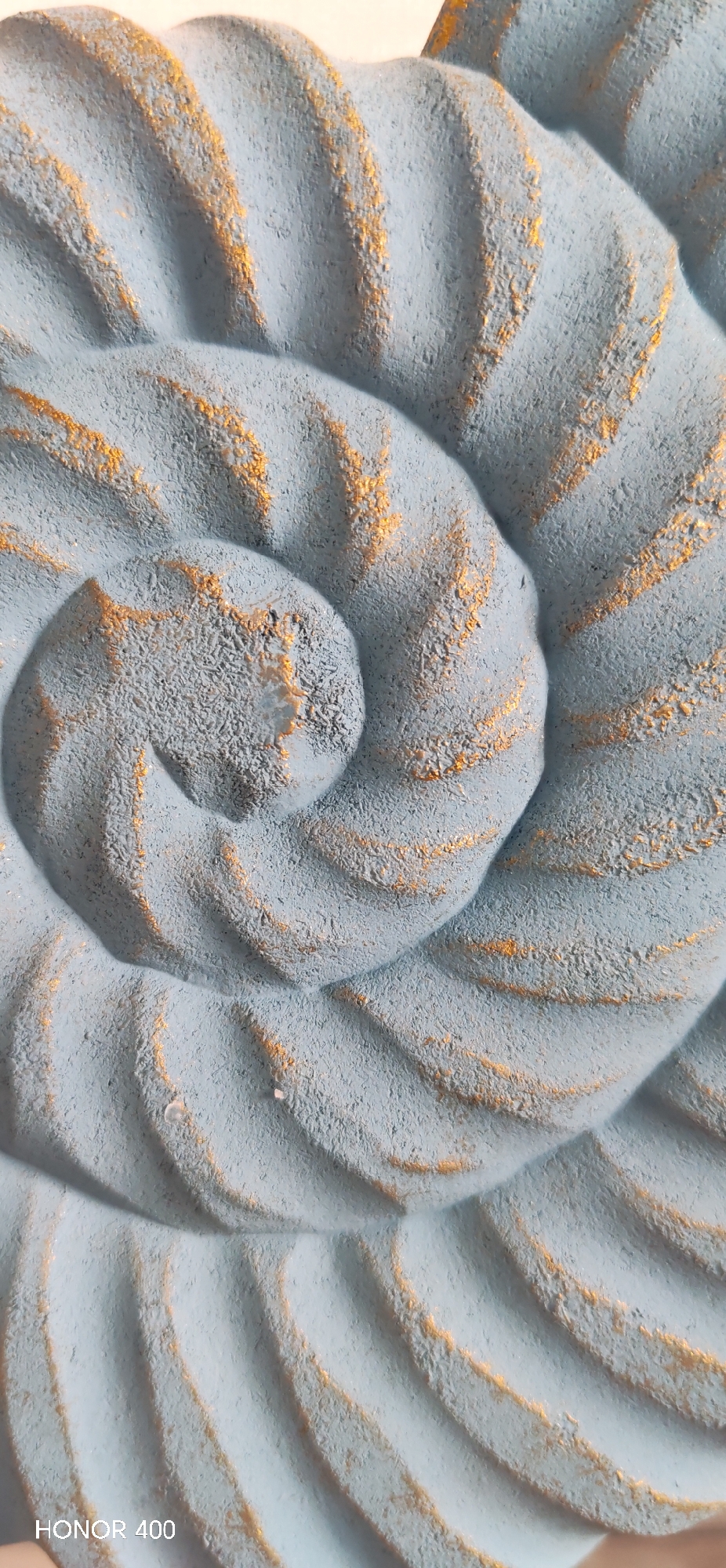 A close-up shot captures a spiral-shaped object. The object is textured, painted in a muted blue-gray, and has traces of gold accentuating its grooves. The bottom left corner of the image shows text that reads "HONOR 400". The absence of any human subjects and contextual setting prevents pinpointing the location to Noviy, Russia.